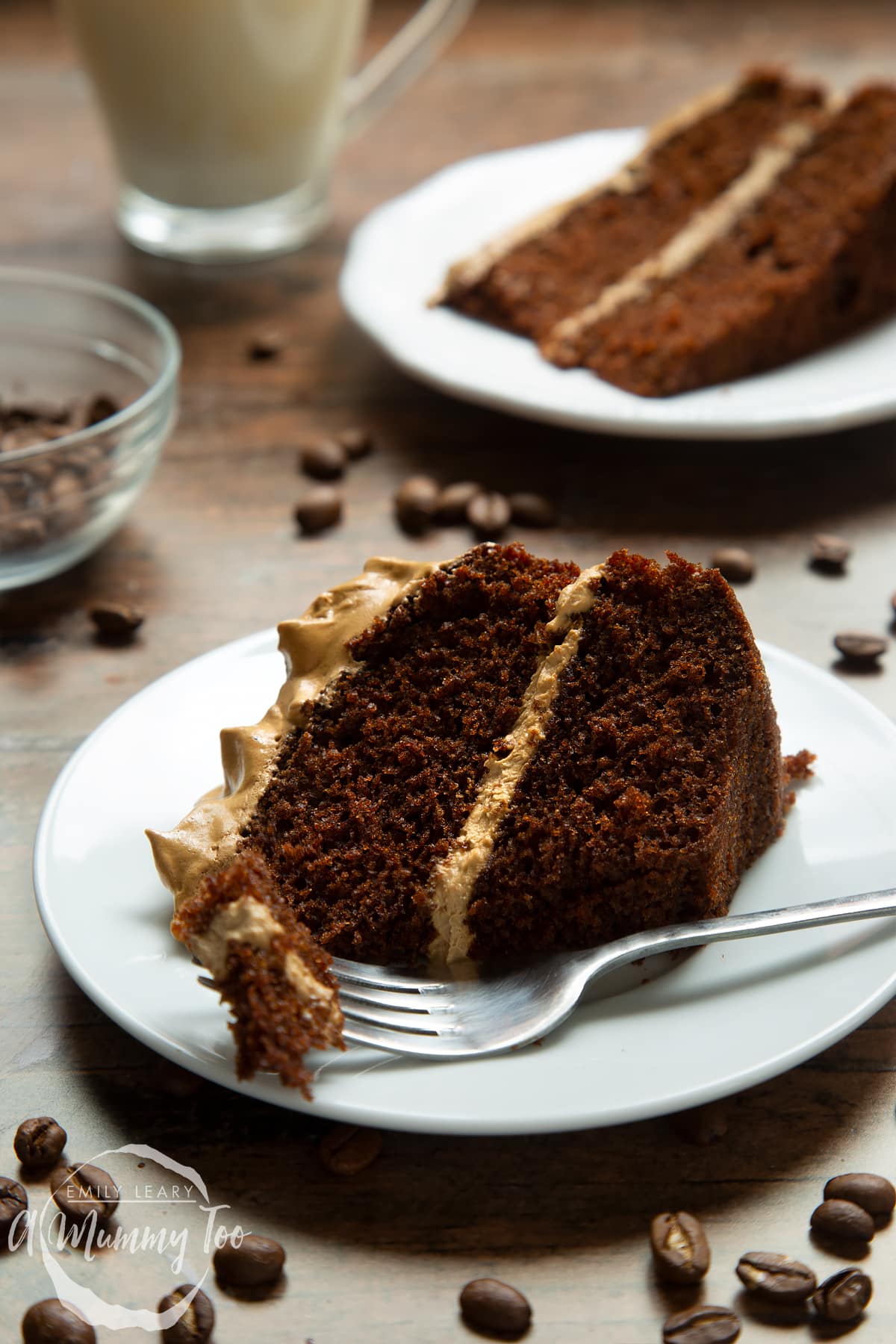 A slice of dalgona coffee cake on a white plate with a fork, which has some cake on. A cup of dalgona coffee, coffee beans and another slice of cake is in the background.