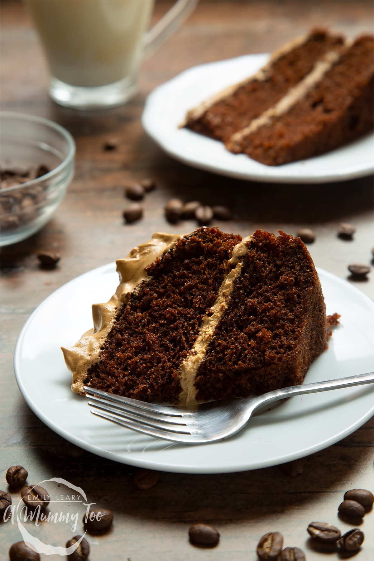 A slice of dalgona coffee cake on a white plate with a fork. A cup of dalgona coffee, coffee beans and another slice of cake is in the background.