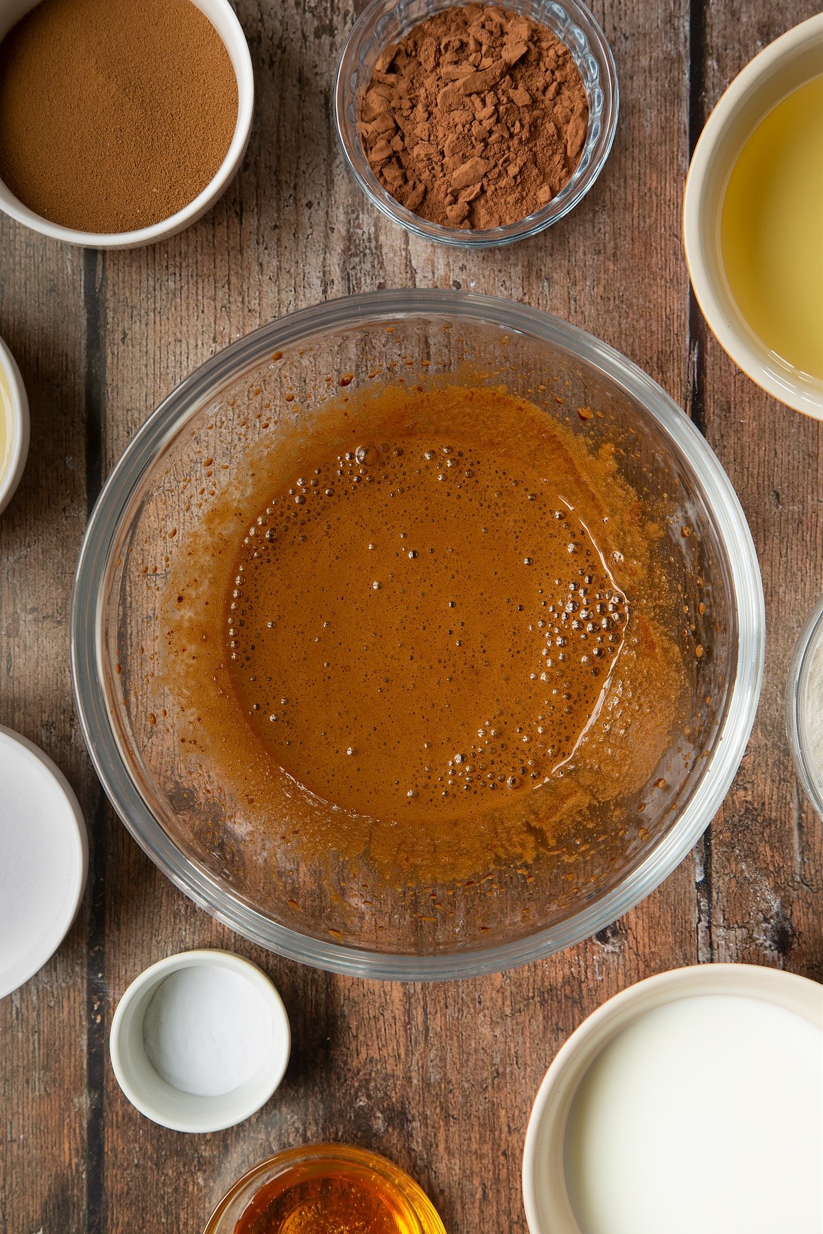 Sugar, instant coffee and hot water mixed together in a glass mixing bowl. Surrounding the bowl is ingredients to make dalgona coffee cake.