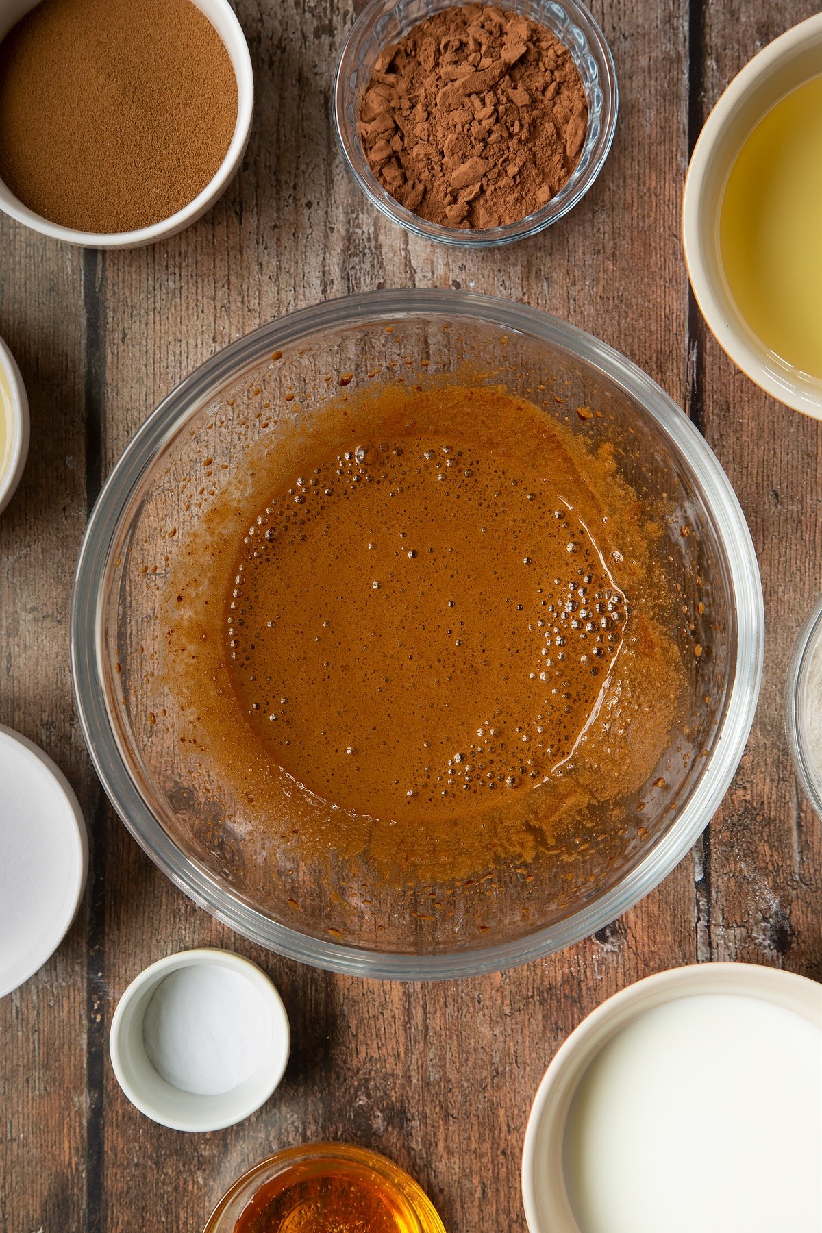 Sucre, café instantané et eau chaude mélangés dans un bol en verre. Autour du bol se trouvent les ingrédients nécessaires à la préparation d'un gâteau au café dalgona.