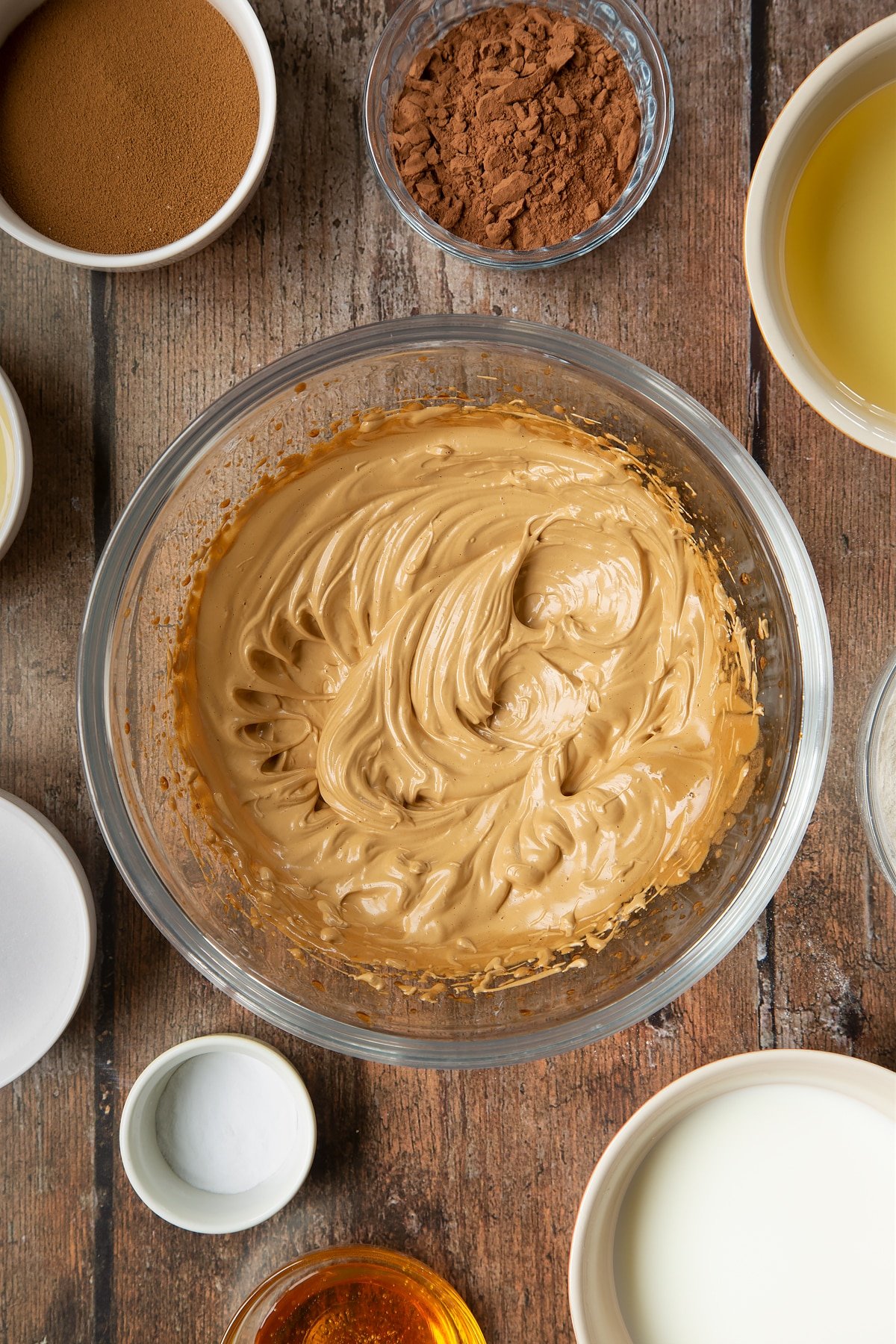 Sugar, instant coffee and hot water whisked together in a glass mixing bowl to create pale, creamy peaks. Surrounding the bowl is ingredients to make dalgona coffee cake.