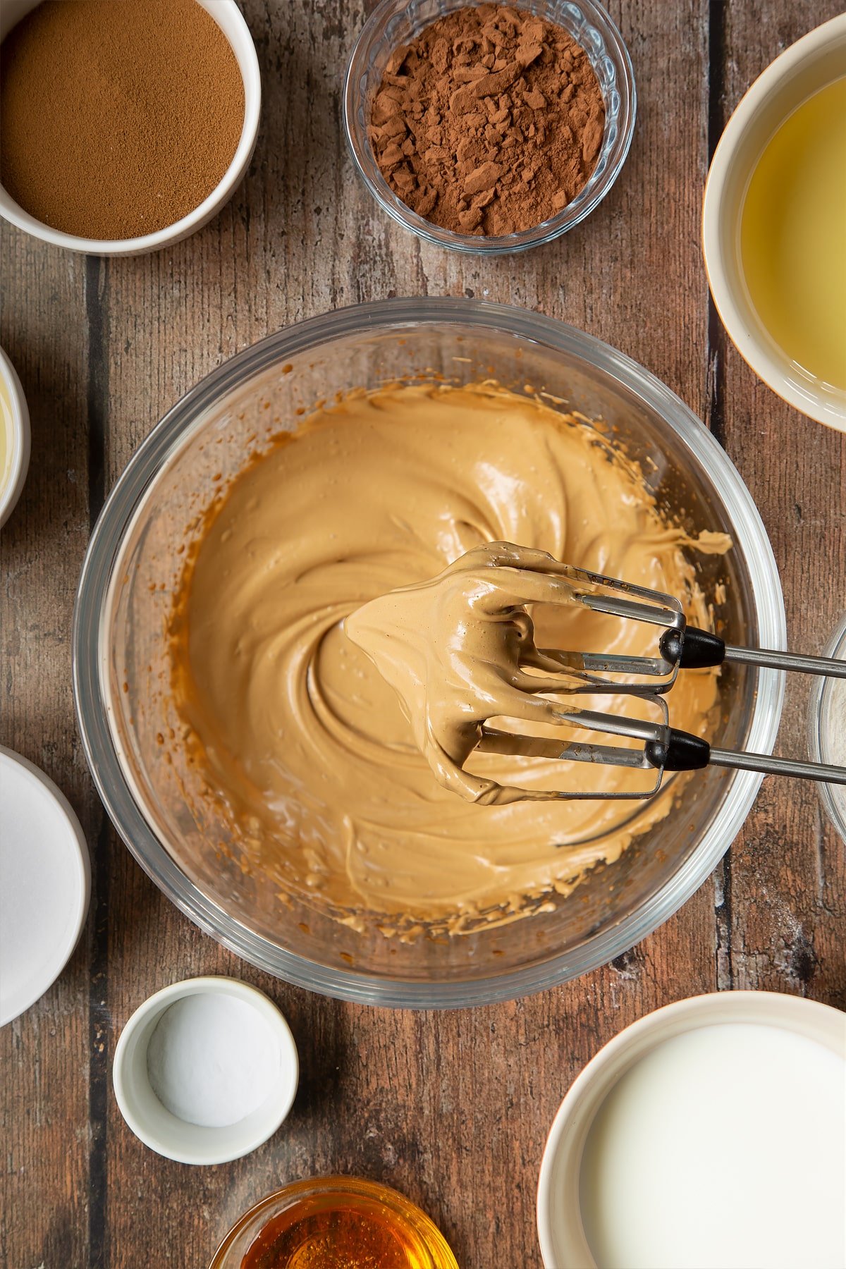 Sugar, instant coffee and hot water whisked together in a glass mixing bowl to create pale, creamy peaks which are shown on the ends of whisk beaters. Surrounding the bowl is ingredients to make dalgona coffee cake.