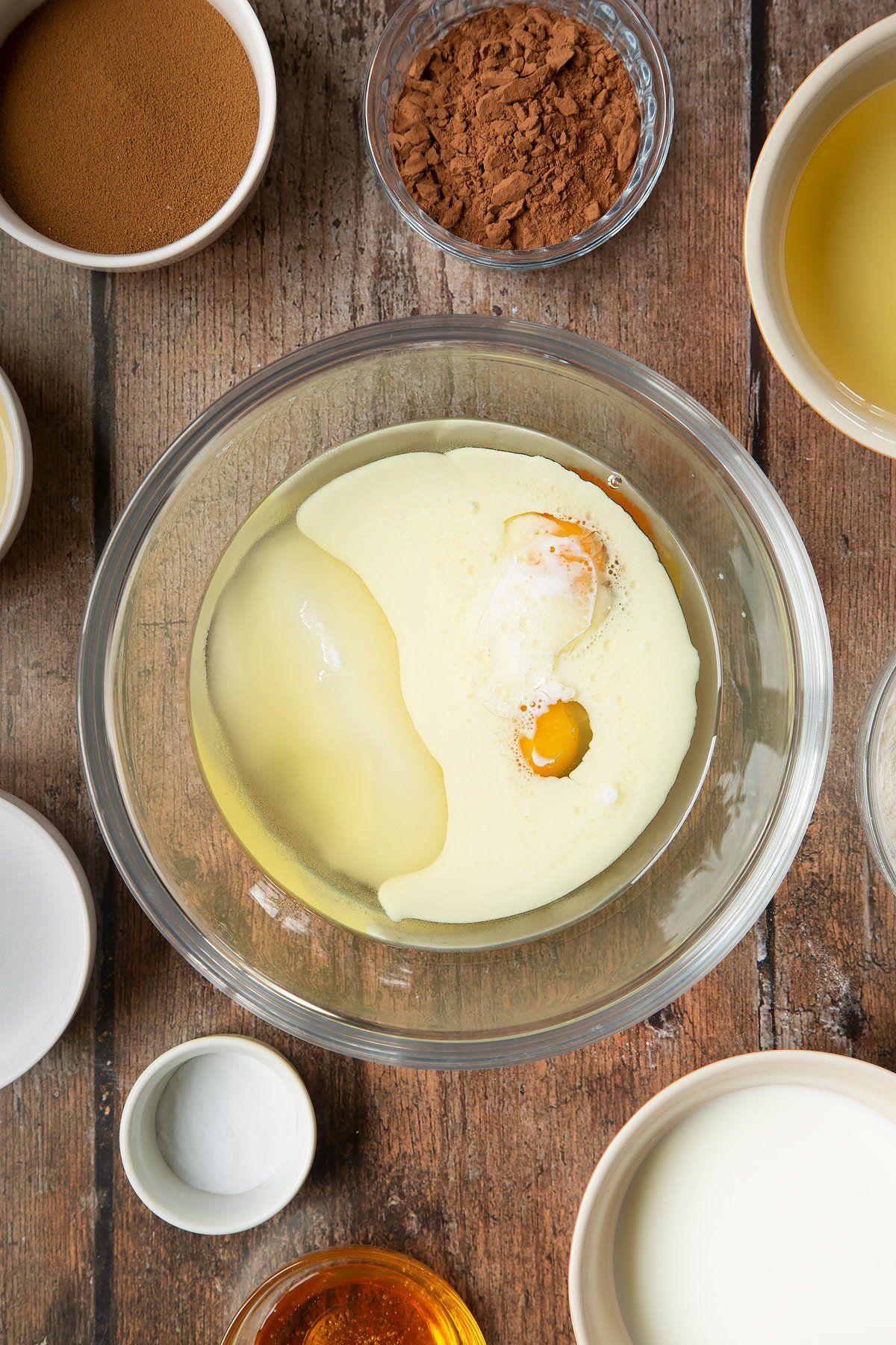 Eggs, sugar, oil, milk and golden syrup in a glass mixing bowl. Surrounding the bowl is ingredients to make dalgona coffee cake.