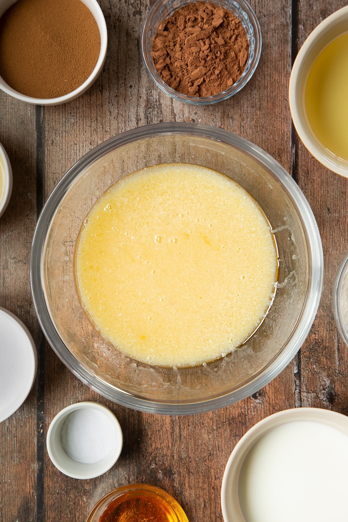 Eggs, sugar, oil, milk and golden syrup whisked together in a glass mixing bowl. Surrounding the bowl is ingredients to make dalgona coffee cake.