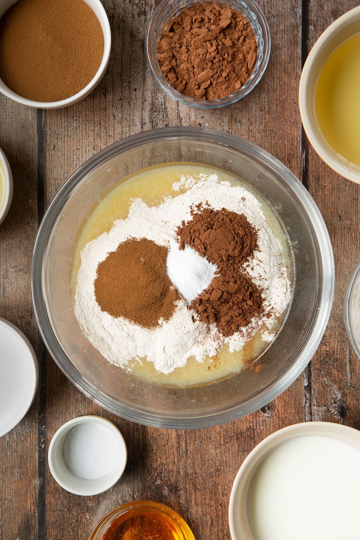 Eggs, sugar, oil, milk and golden syrup whisked together in a glass mixing bowl with flour, cocoa, instant coffee and bicarbonate of soda on top. Surrounding the bowl is ingredients to make dalgona coffee cake.