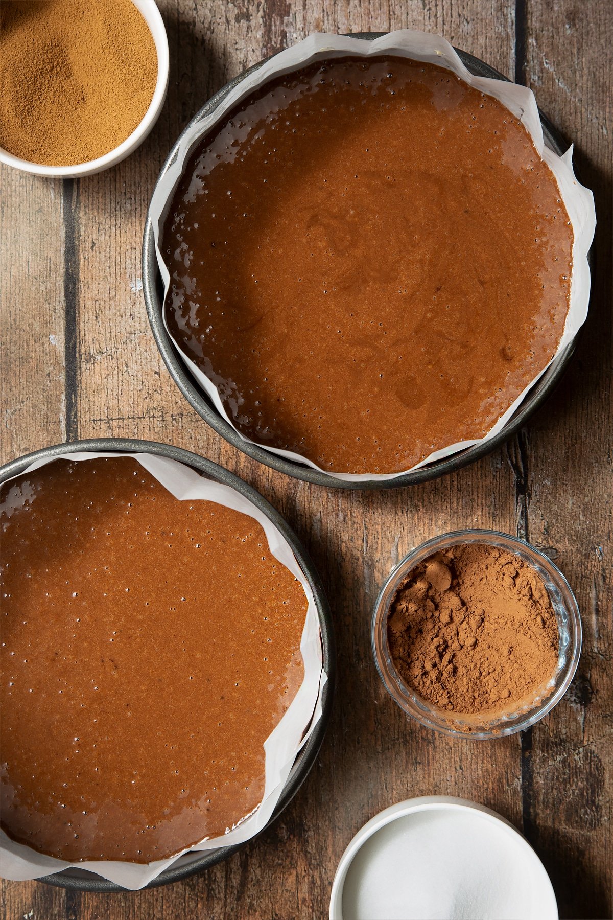 Cake mix in two lined cake tins. Surrounding the bowl is ingredients to make dalgona coffee cake.