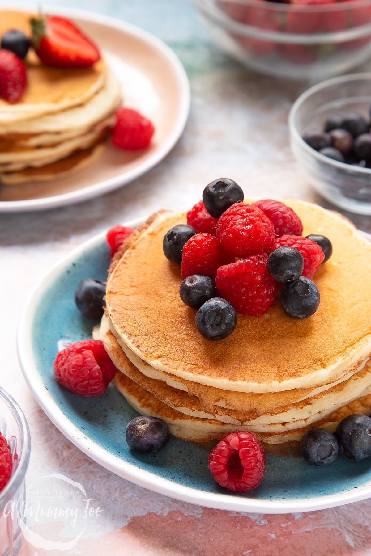 Stack of skyr pancakes on a blue plate. The pancakes are topped with blueberries and raspberries. Another stack of pancakes is shown on a pink plate in the background.