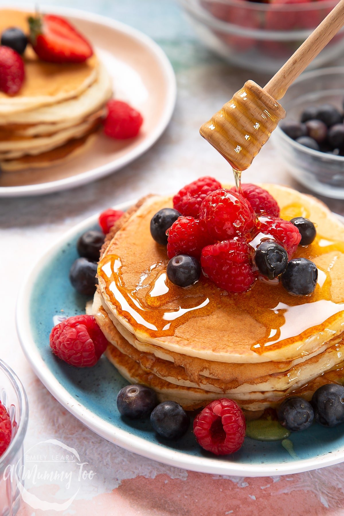 Stack of skyr pancakes on a blue plate. The pancakes are topped with blueberries and raspberries. A honey stick drizzles honey over the pancakes.