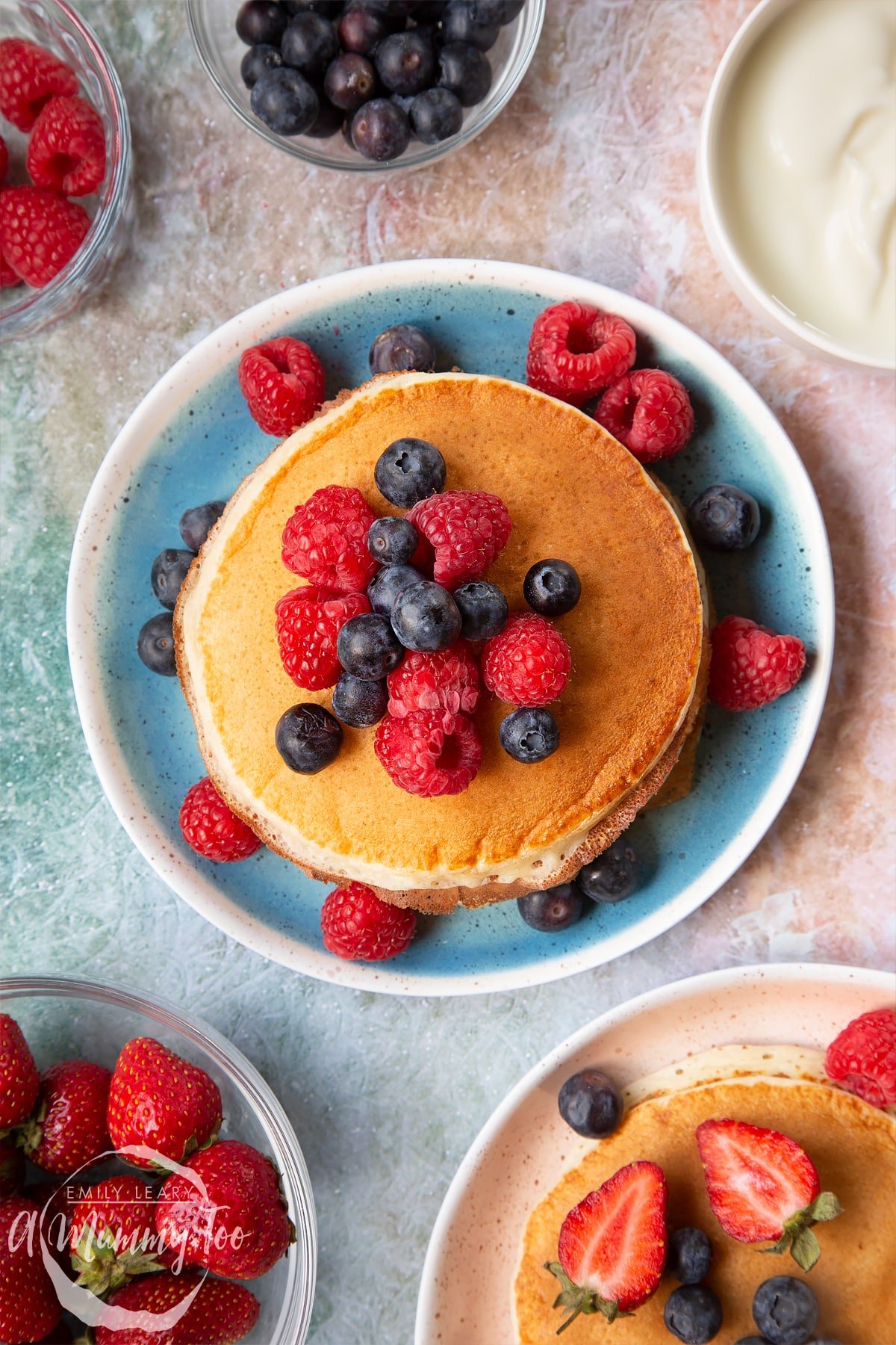 Stack of skyr pancakes on a blue plate. The pancakes are topped with blueberries and raspberries. Bowls of berries surround the plate.