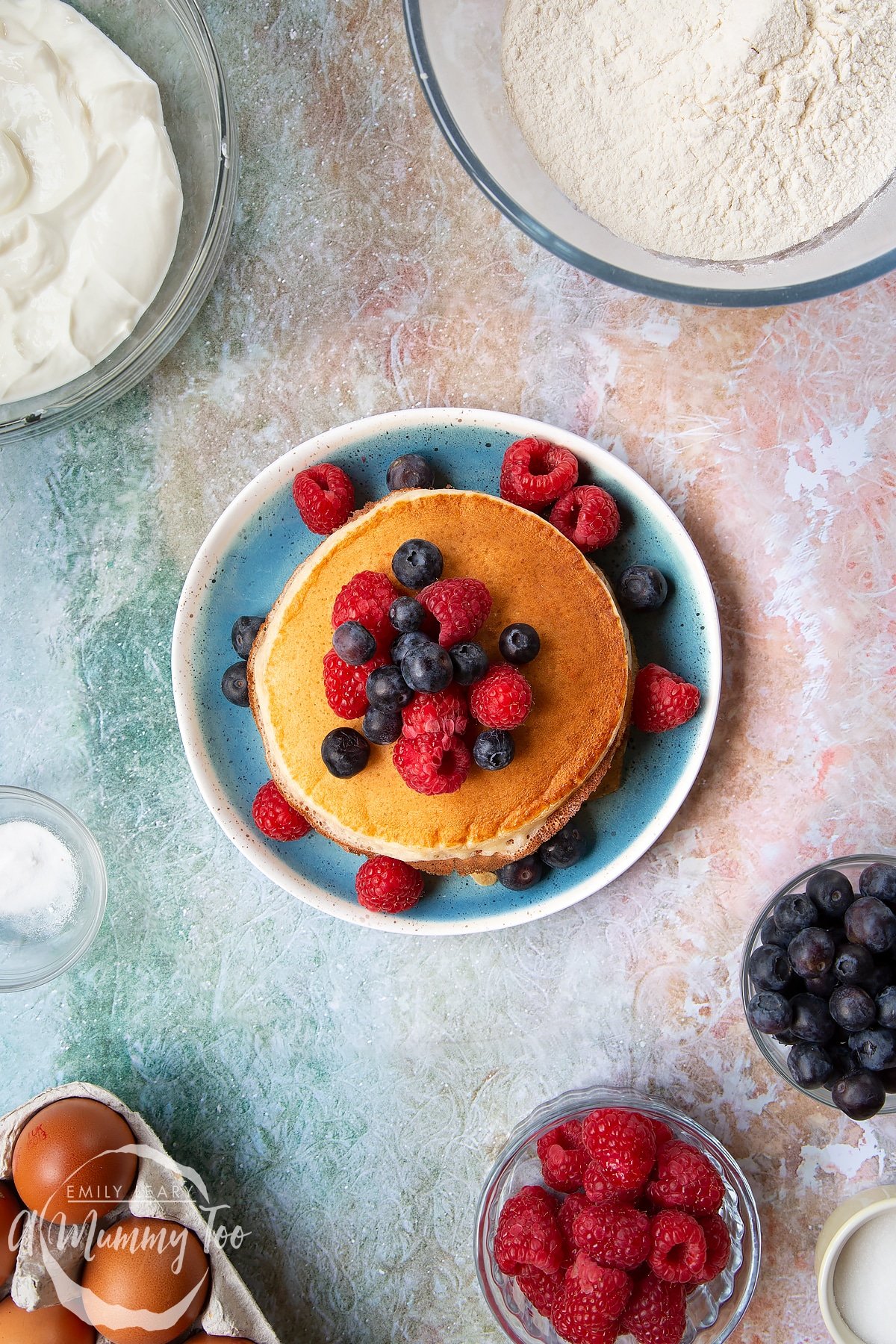 Skyr pancakes piled on a blue plate with blueberries and raspberries on top. Berries and ingredients to make skyr pancakes surround the pan.