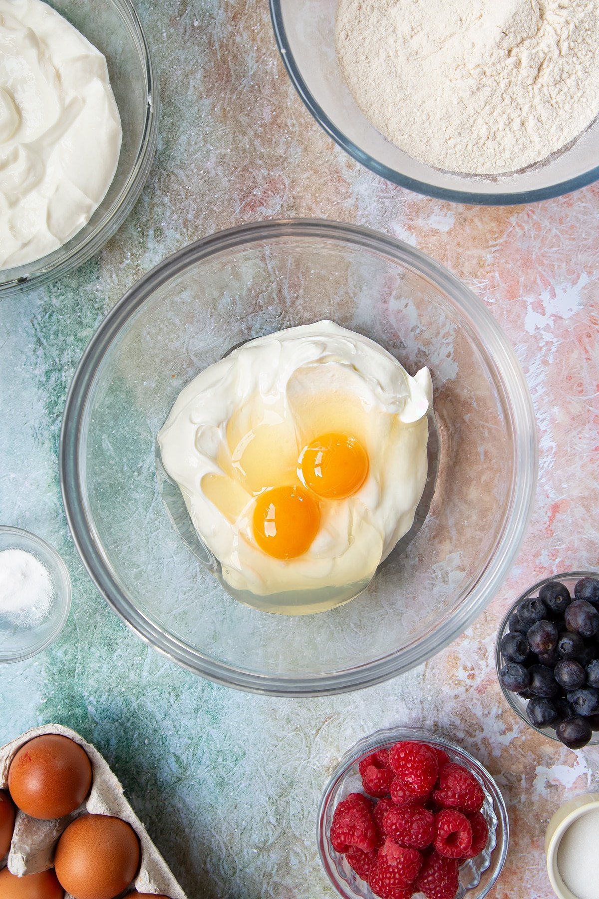 Eggs and skyr in a mixing bowl. Berries and ingredients to make skyr pancakes surround the bowl.