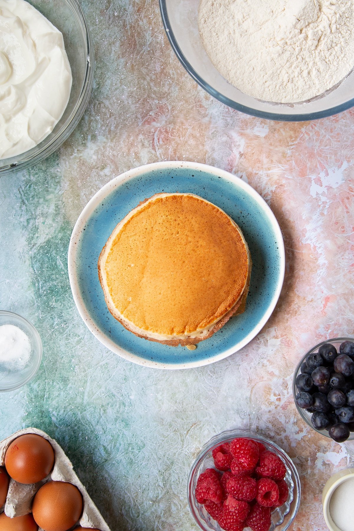 Skyr pancakes piled on a blue plate. Berries and ingredients to make skyr pancakes surround the pan.