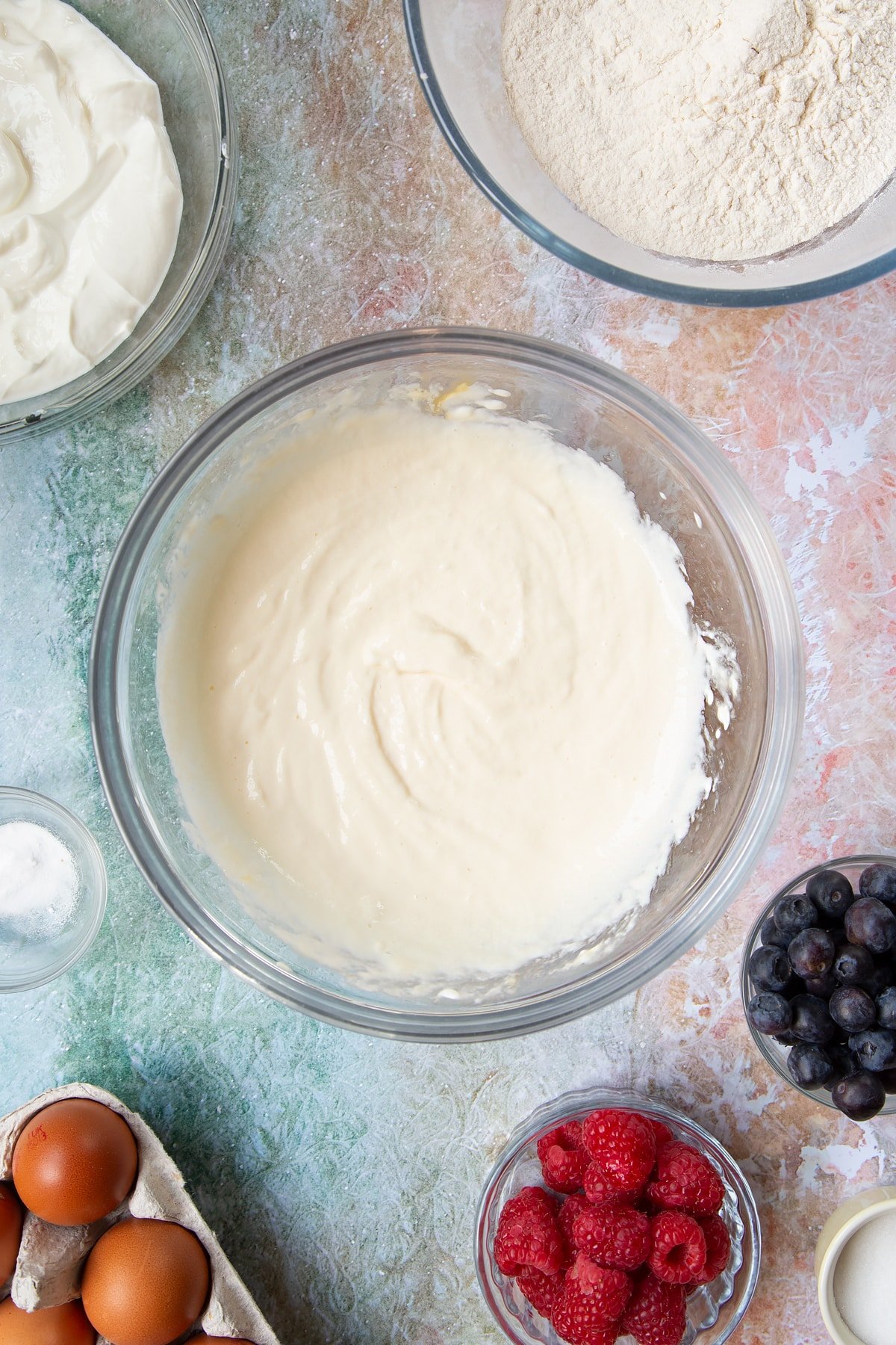 Eggs, skyr and milk whisked together in a mixing bowl. Berries and ingredients to make skyr pancakes surround the bowl.