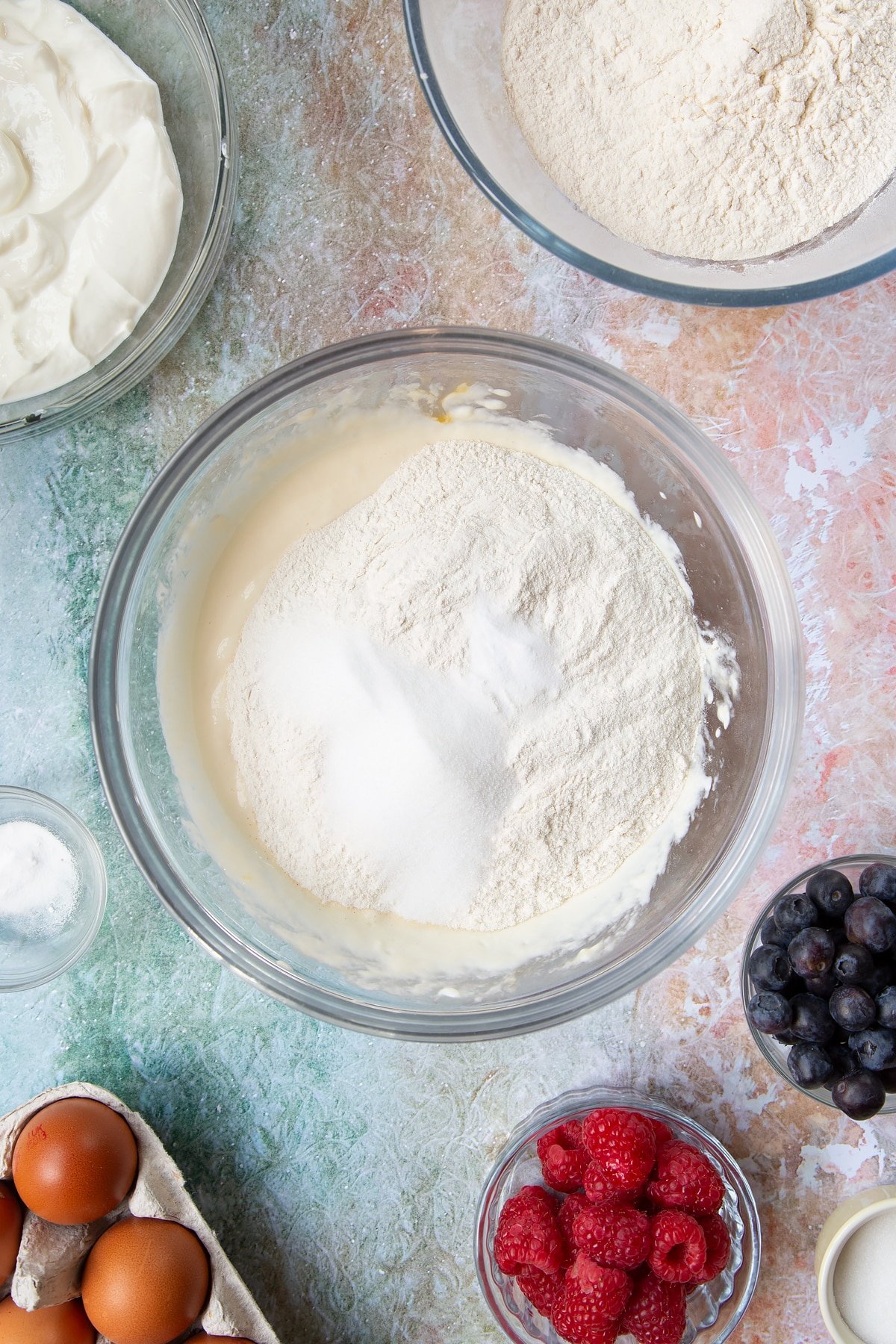 Eggs, skyr and milk whisked together in a mixing bowl with flour, baking powder, baking soda and sugar on top. Berries and ingredients to make skyr pancakes surround the bowl.