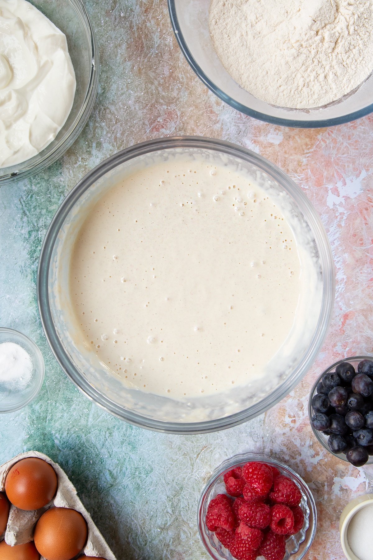 Skyr pancake batter in a mixing bowl. Berries and ingredients to make skyr pancakes surround the bowl.