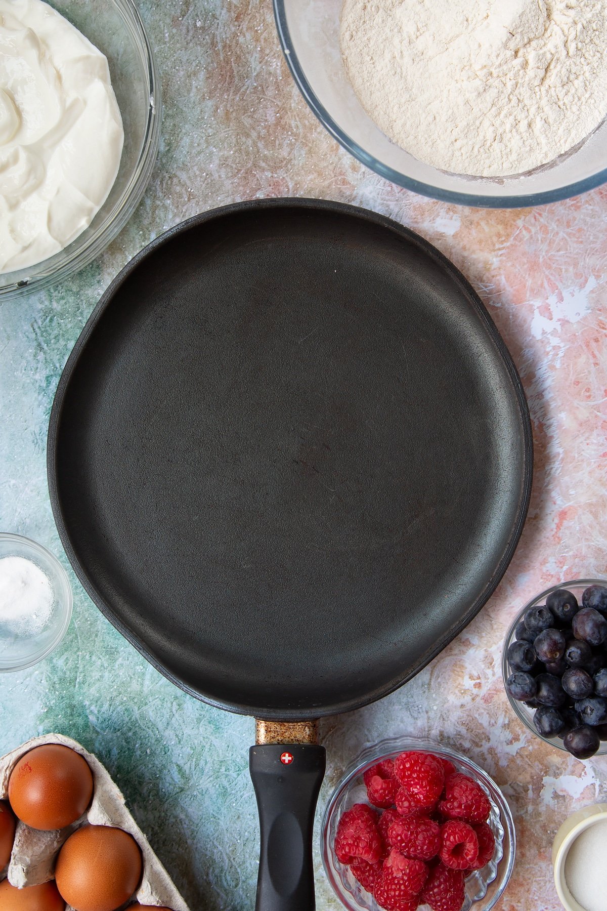 A hot frying pan. Berries and ingredients to make skyr pancakes surround the pan.