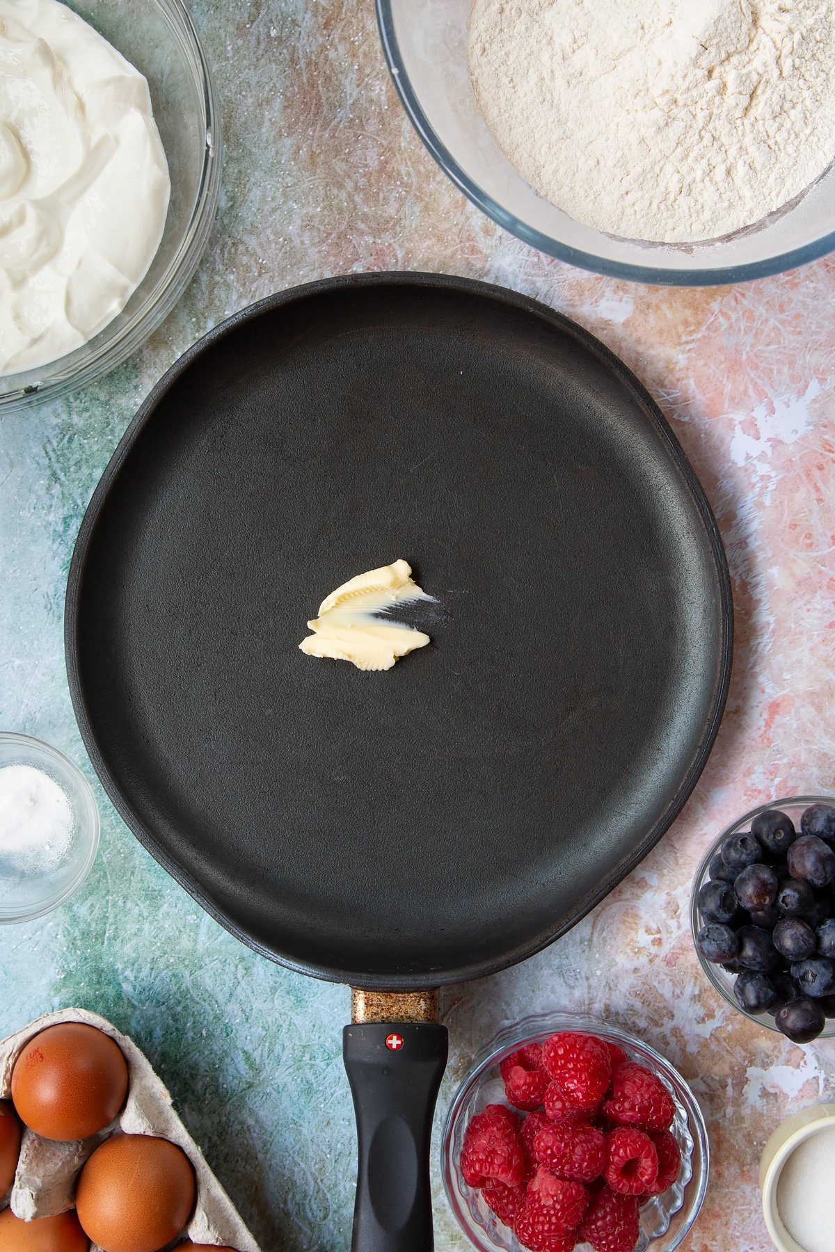 A hot frying pan with butter melting in it. Berries and ingredients to make skyr pancakes surround the pan.