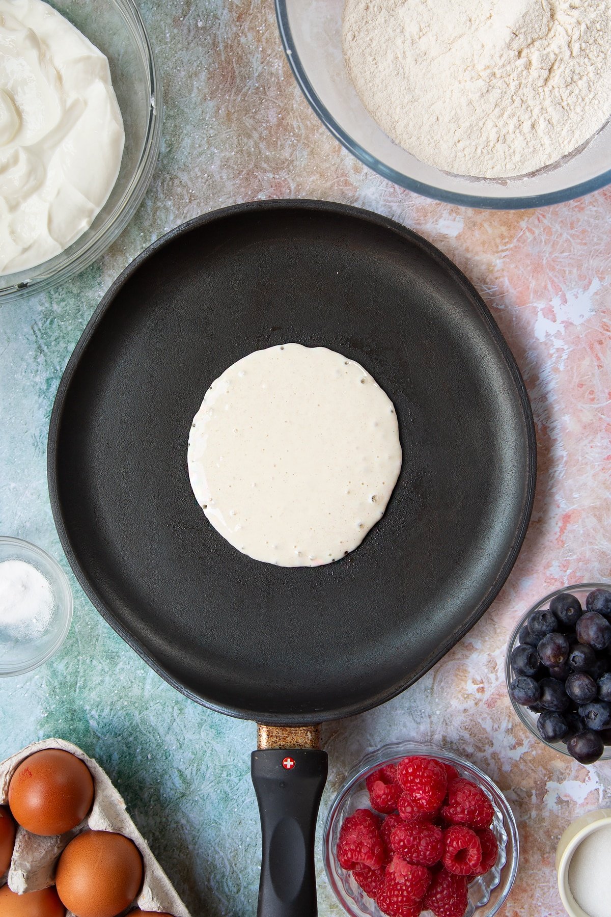 A hot frying pan with a circle of pancake batter in it. Berries and ingredients to make skyr pancakes surround the pan.