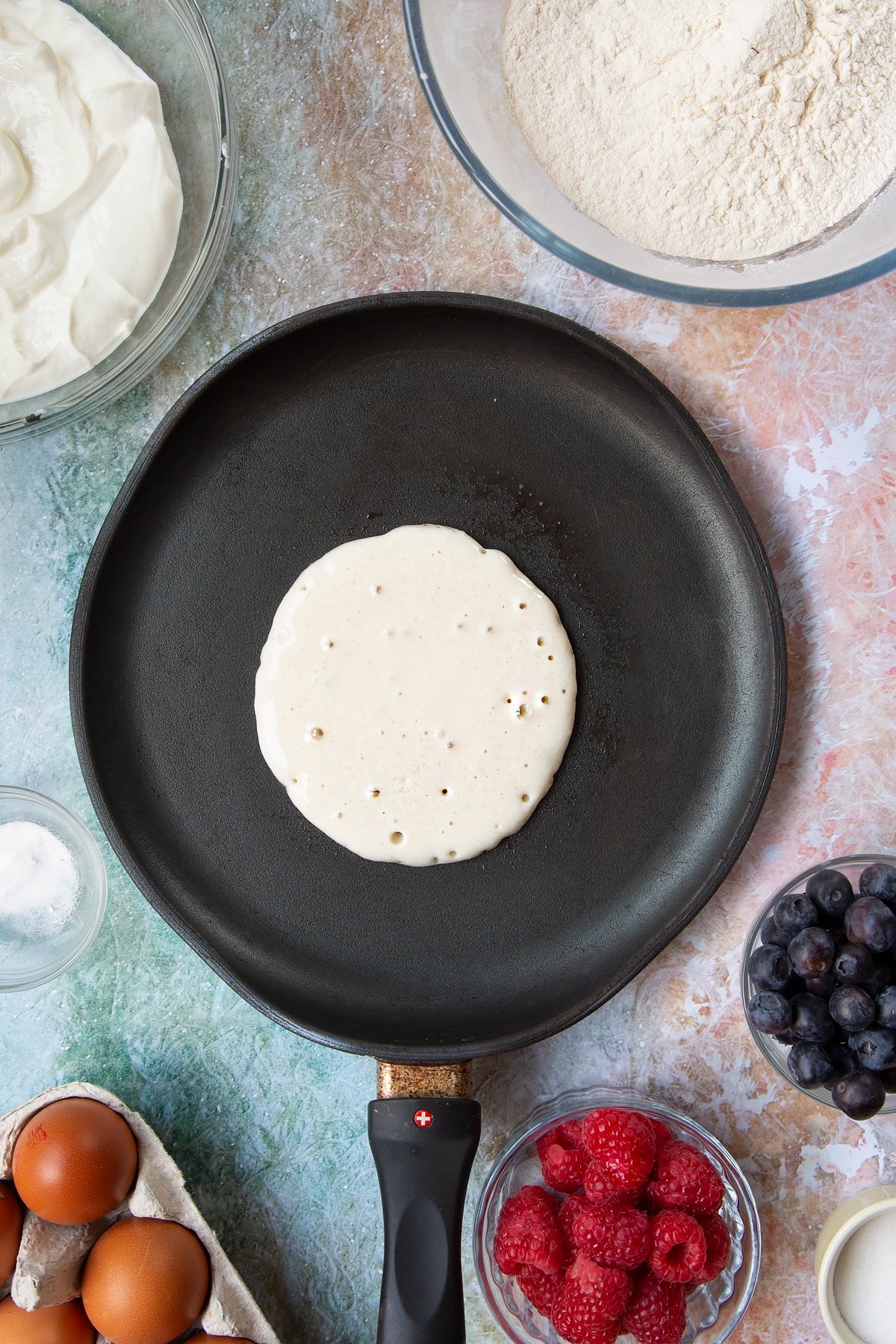 A hot frying pan with a circle of pancake batter in it, which is forming bubbles on the surface. Berries and ingredients to make skyr pancakes surround the pan.