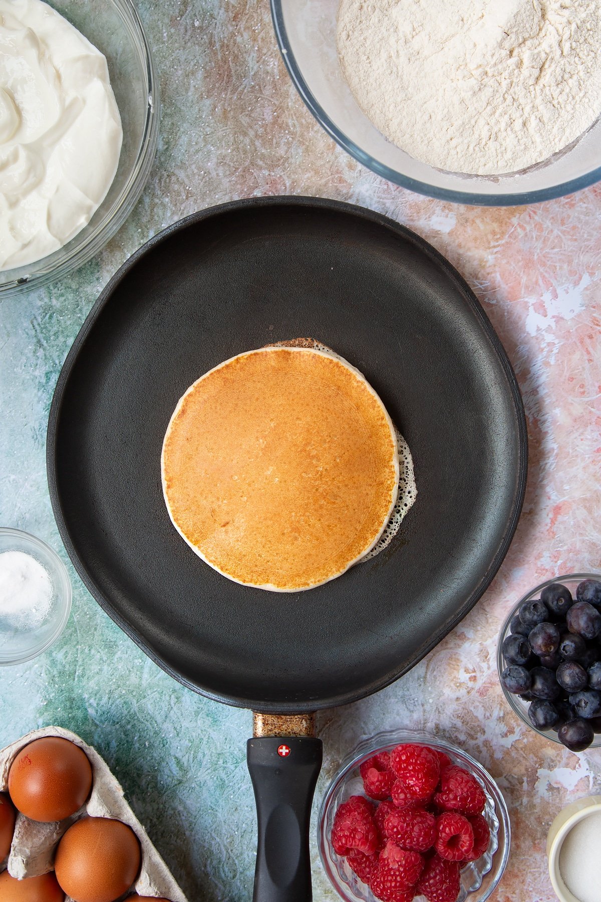 A hot frying pan with a cooked skyr pancake in it. Berries and ingredients to make skyr pancakes surround the pan.