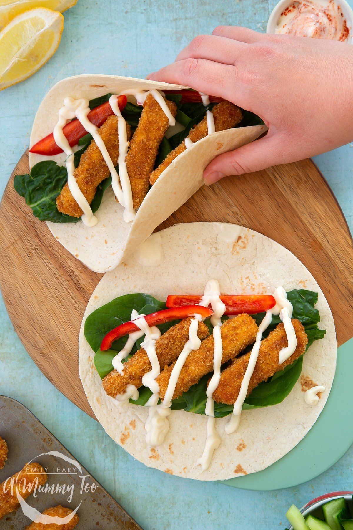 Hand picking up a flour tortilla filled with tofu fish sticks, salad and mayo. Another open tortilla with the same filling is beside it on a wooden board.