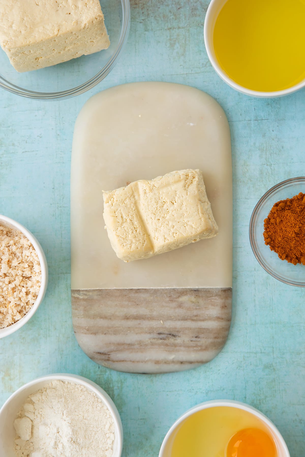 A block of firm tofu on a small marble board. Ingredients to make tofu fingers surround the board.