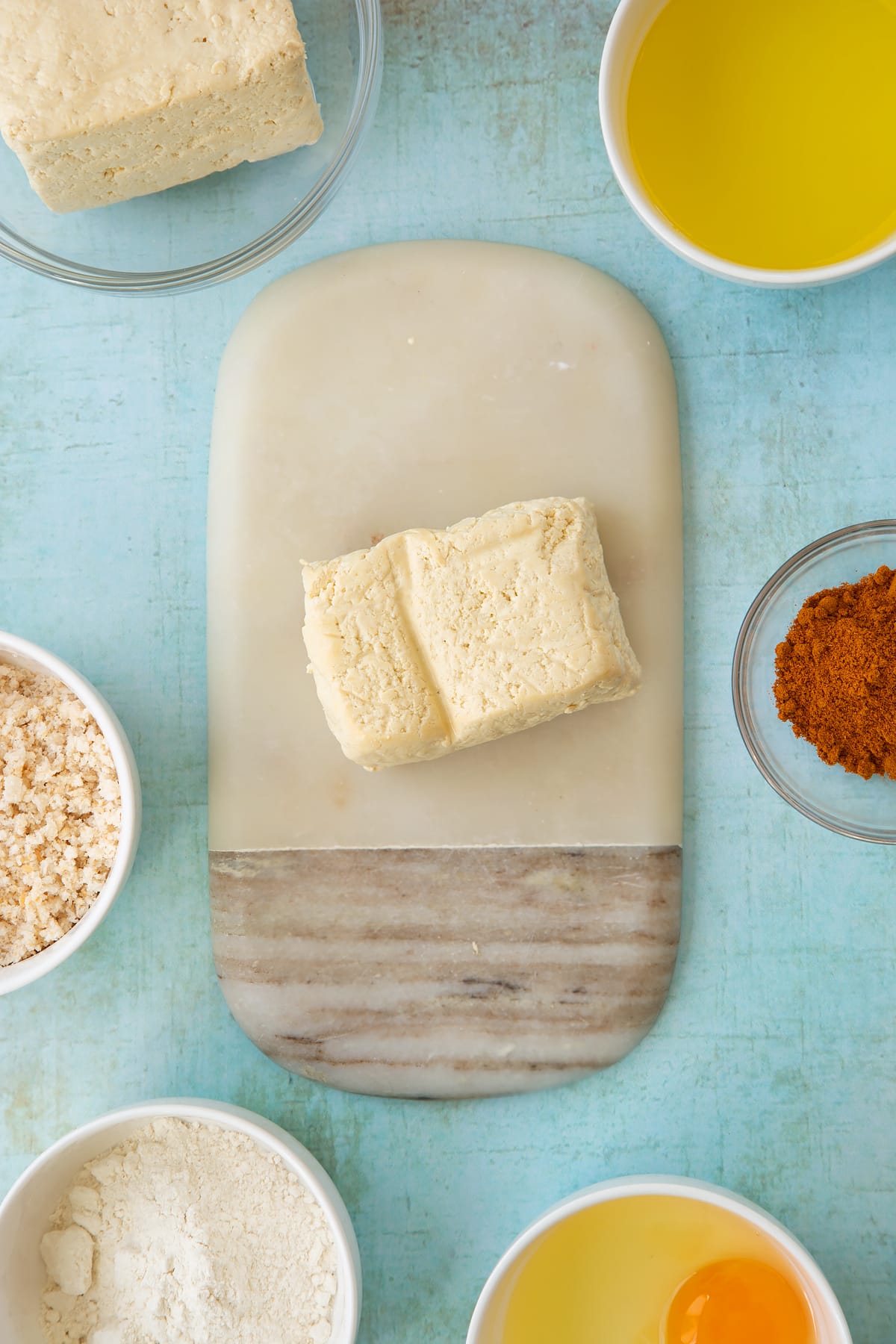 A block of firm tofu on a small marble board. Ingredients to make tofu fish sticks surround the board.