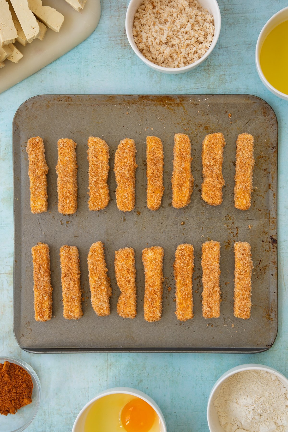 Uncooked, breadcrumb coated tofu fish sticks on a baking sheet. Ingredients to make tofu fish sticks surround the sheet.