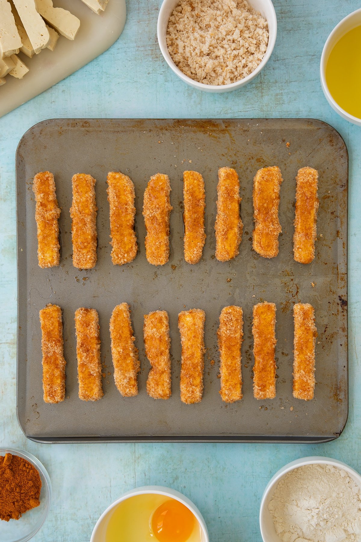 Uncooked, breadcrumb coated tofu fish sticks on a baking sheet, drizzled with oil. Ingredients to make tofu fish sticks surround the sheet.