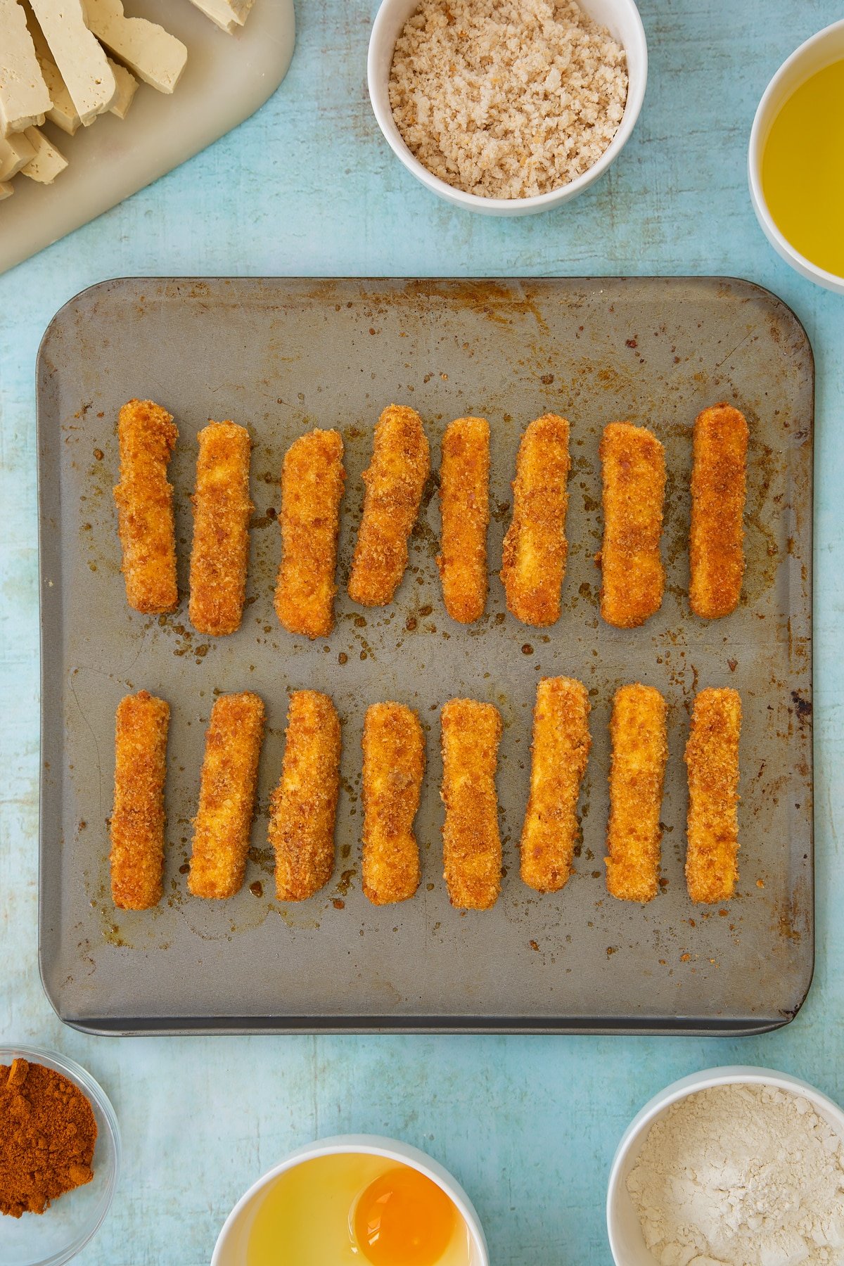 Freshly baked, breadcrumb coated tofu fish sticks on a baking sheet. Ingredients to make tofu fish sticks surround the sheet.