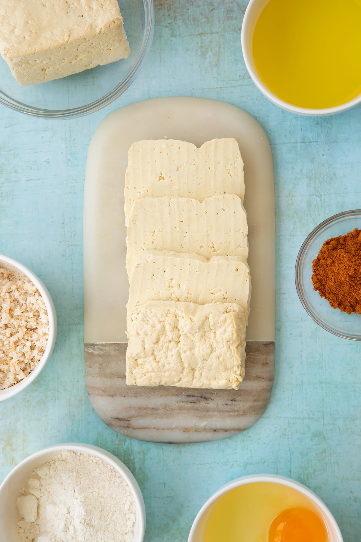 A block of firm tofu cut into slabs on a small marble board. Ingredients to make tofu fish sticks surround the board.