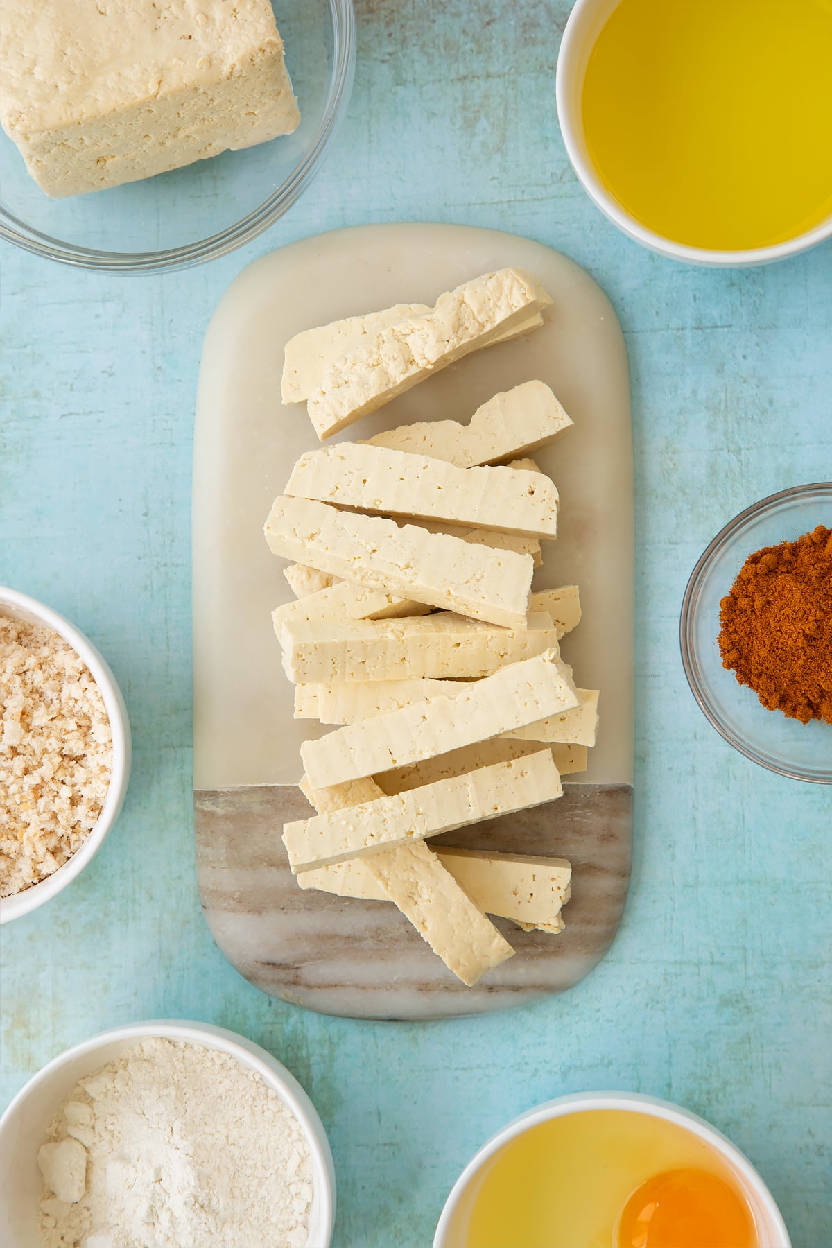 A block of firm tofu cut into fingers on a small marble board. Ingredients to make tofu fingers surround the board.