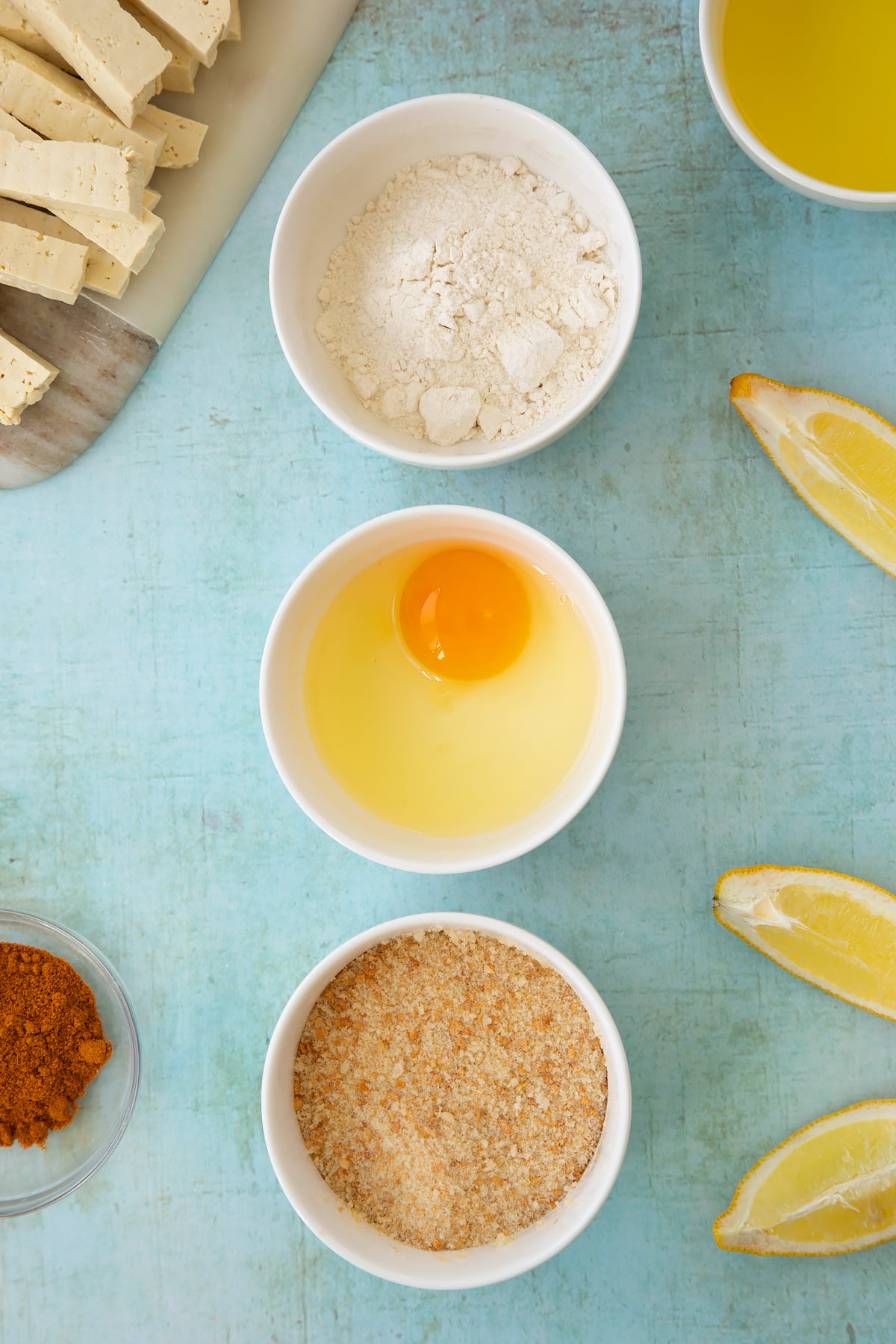 Three small white bowls containing flour, an egg and breadcrumbs. Ingredients to make tofu fish sticks surround the bowls.