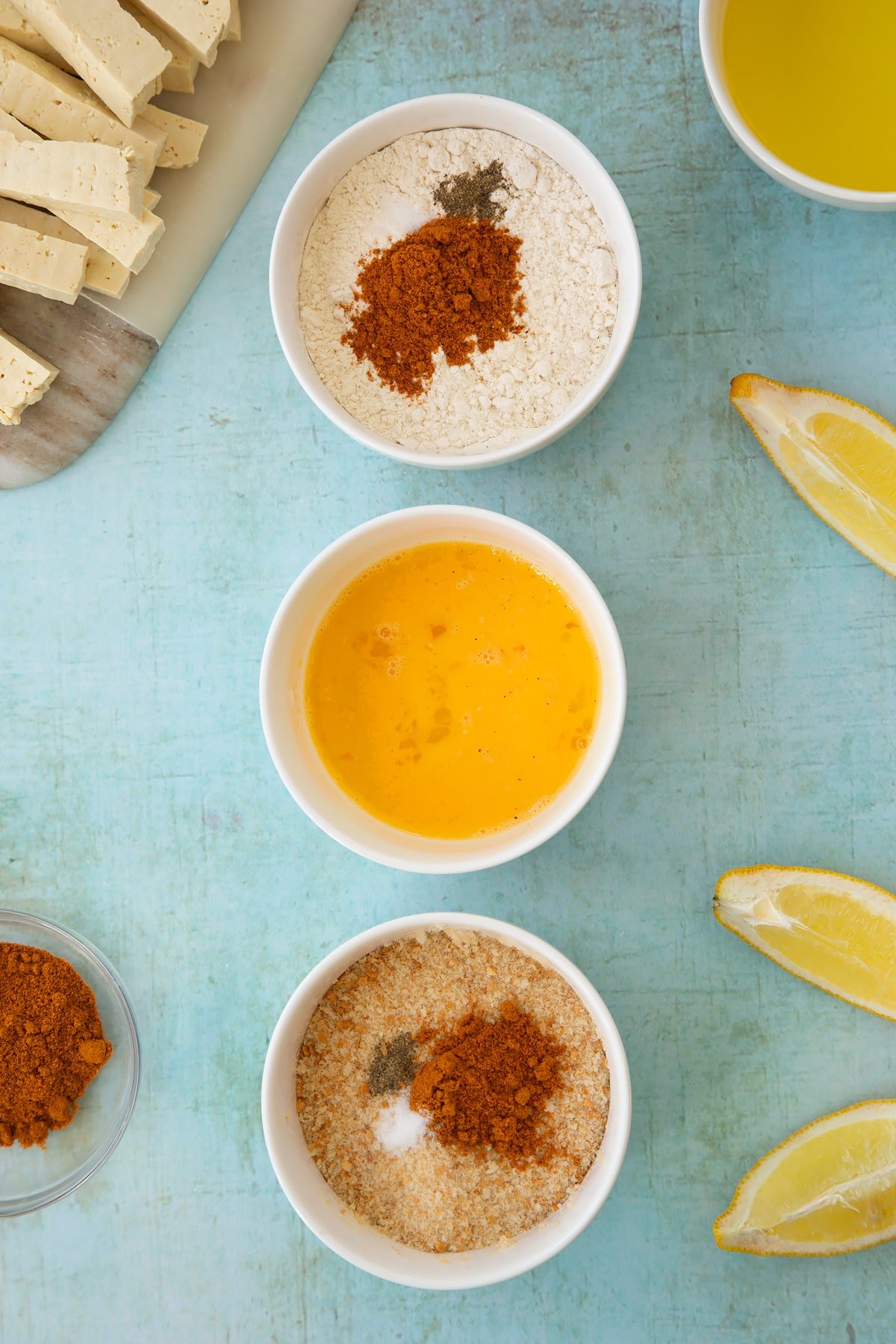 Three small white bowls. The first contains flour with madras powder, the second contains a beaten egg and the third contains breadcrumbs and madras powder. Ingredients to make tofu fish sticks surround the bowls.