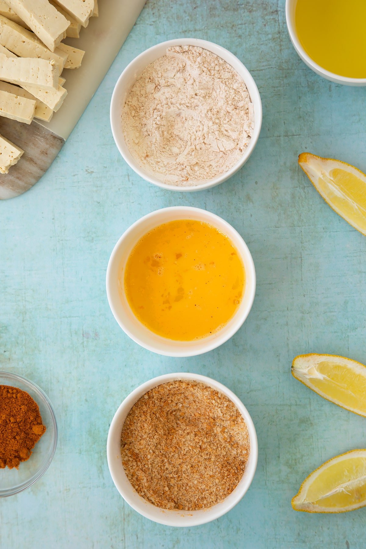 Three small white bowls. The first contains flour mixed with madras powder, the second contains a beaten egg and the third contains breadcrumbs mixed with madras powder. Ingredients to make tofu fish sticks surround the bowls.