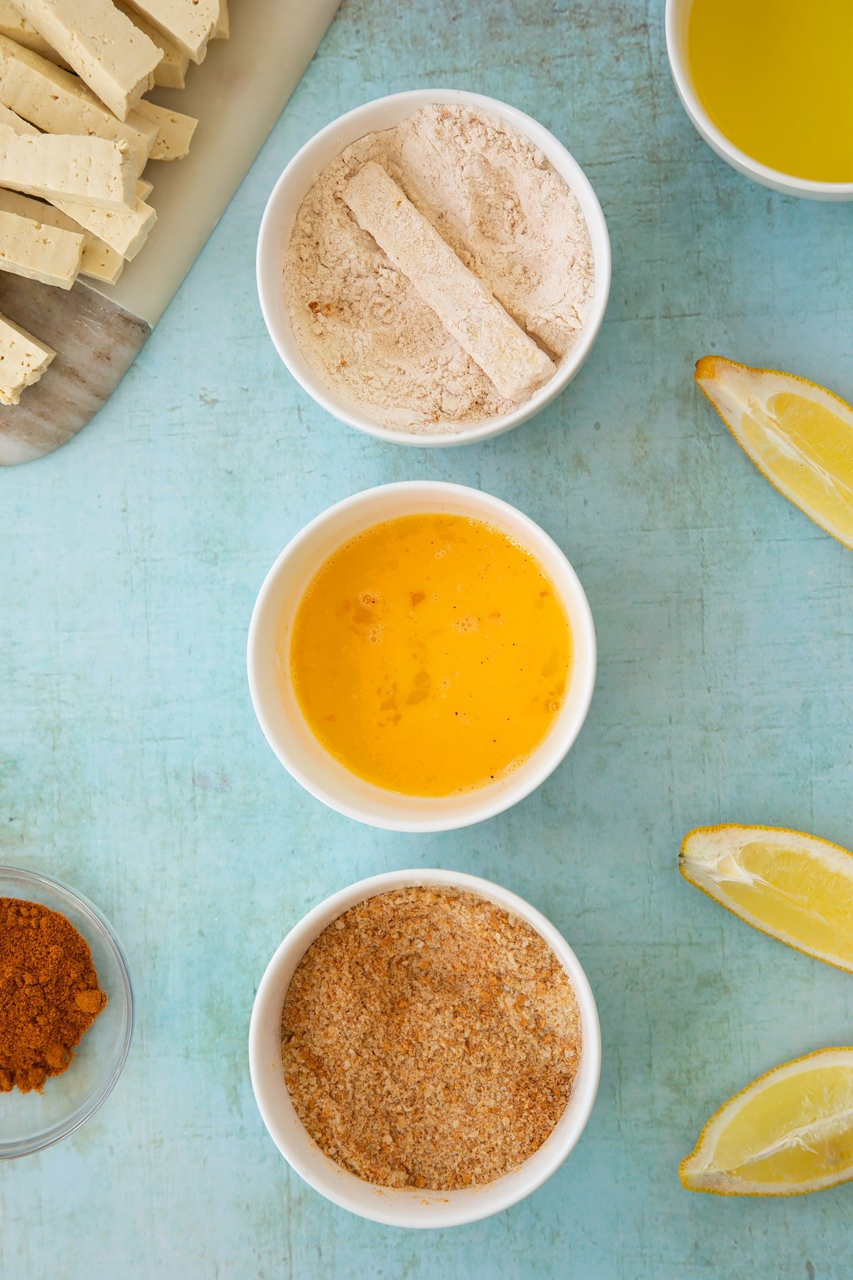 Three small white bowls. The first contains flour mixed with madras powder - a piece of tofu is in the bowl, coated in flour. The second contains a beaten egg and the third contains breadcrumbs mixed with madras powder. Ingredients to make tofu fish sticks surround the bowls.