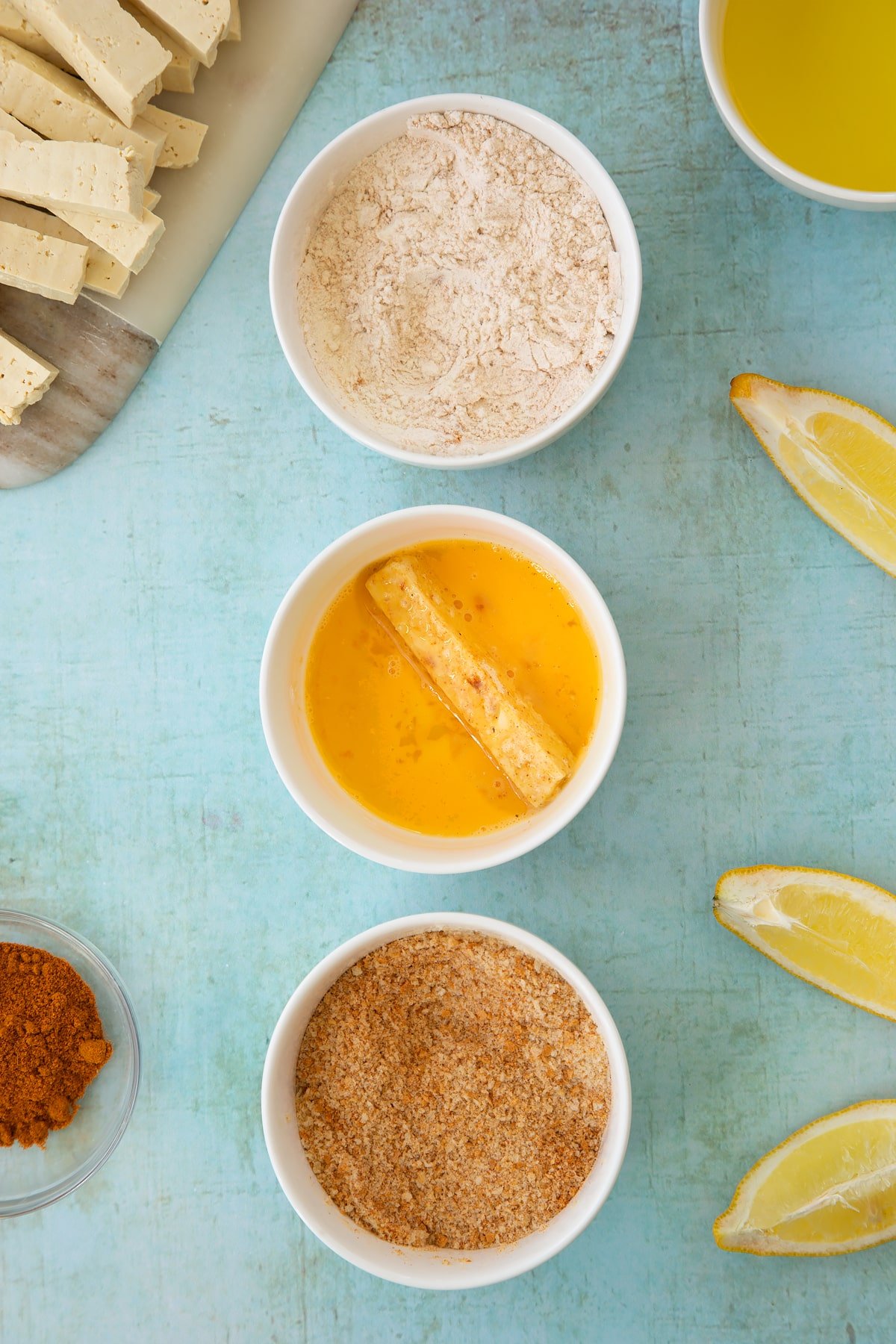 Three small white bowls. The first contains flour mixed with madras powder. The second contains a beaten egg and a piece of tofu, coated in egg. The third contains breadcrumbs mixed with madras powder. Ingredients to make tofu fish sticks surround the bowls.