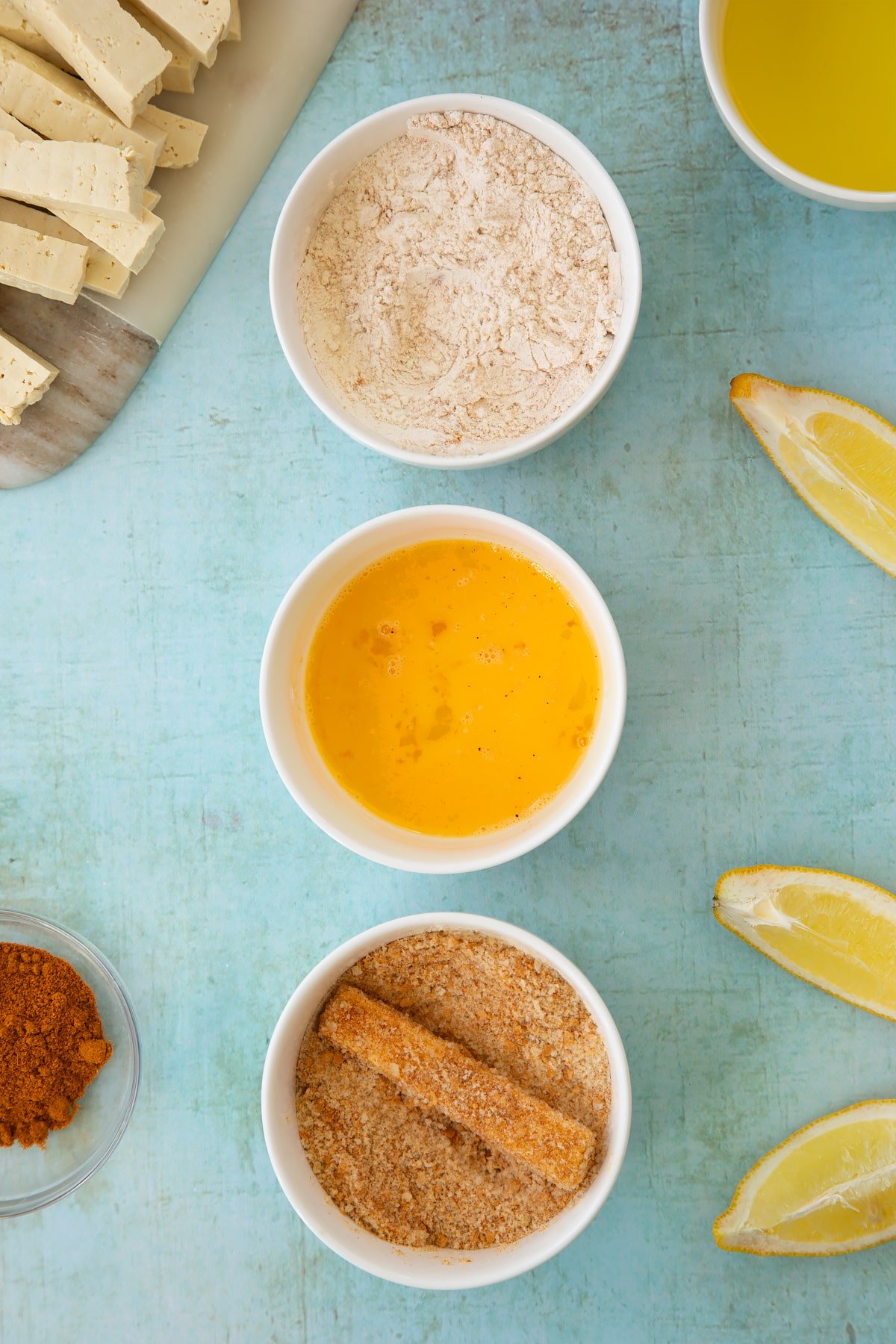 Three small white bowls. The first contains flour mixed with madras powder. The second contains a beaten egg. The third contains breadcrumbs mixed with madras powder - a piece of tofu is also in the bowl, coated with breadcrumbs. Ingredients to make tofu fish sticks surround the bowls.