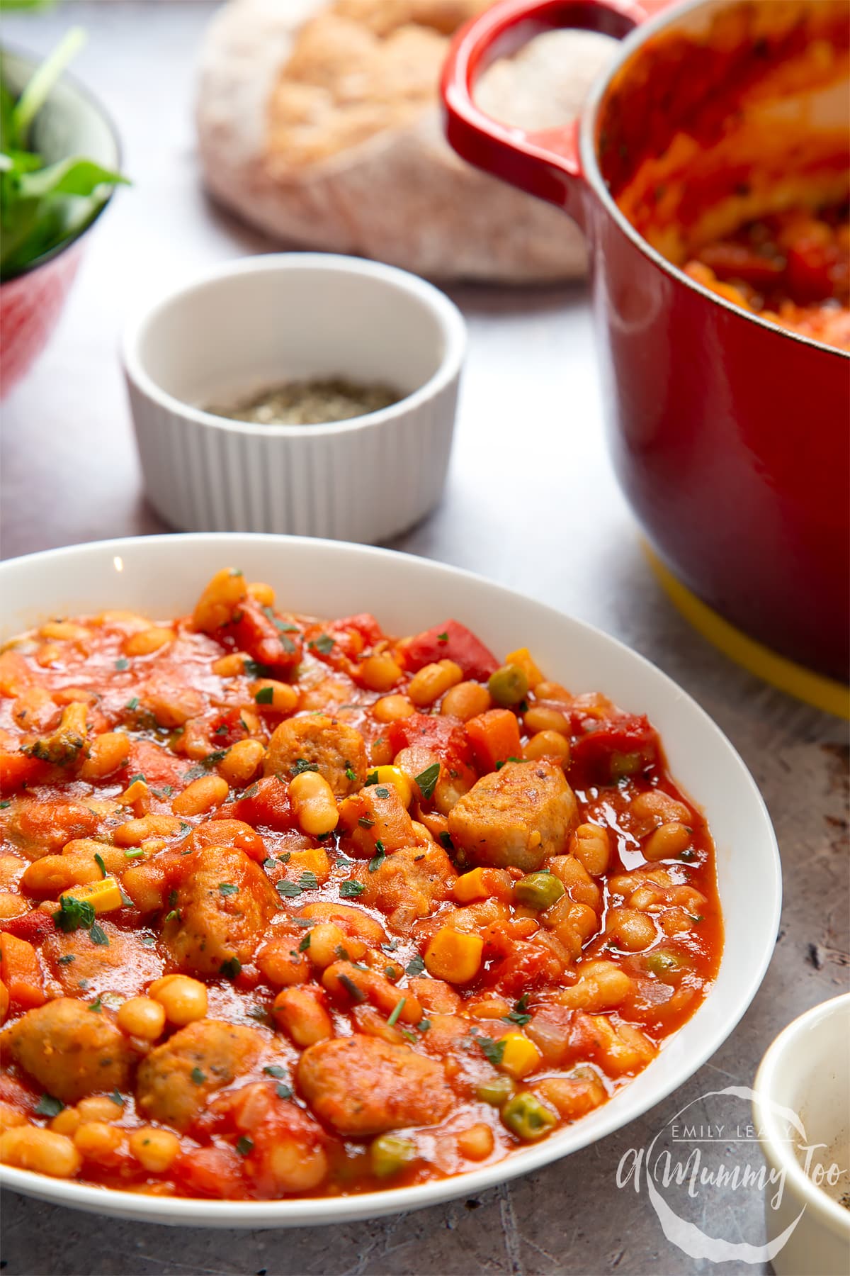 Veggie sausage and bean casserole served into a shallow white bowl. Crusty break, seasoning, salad and a casserole pot are shown in the background.