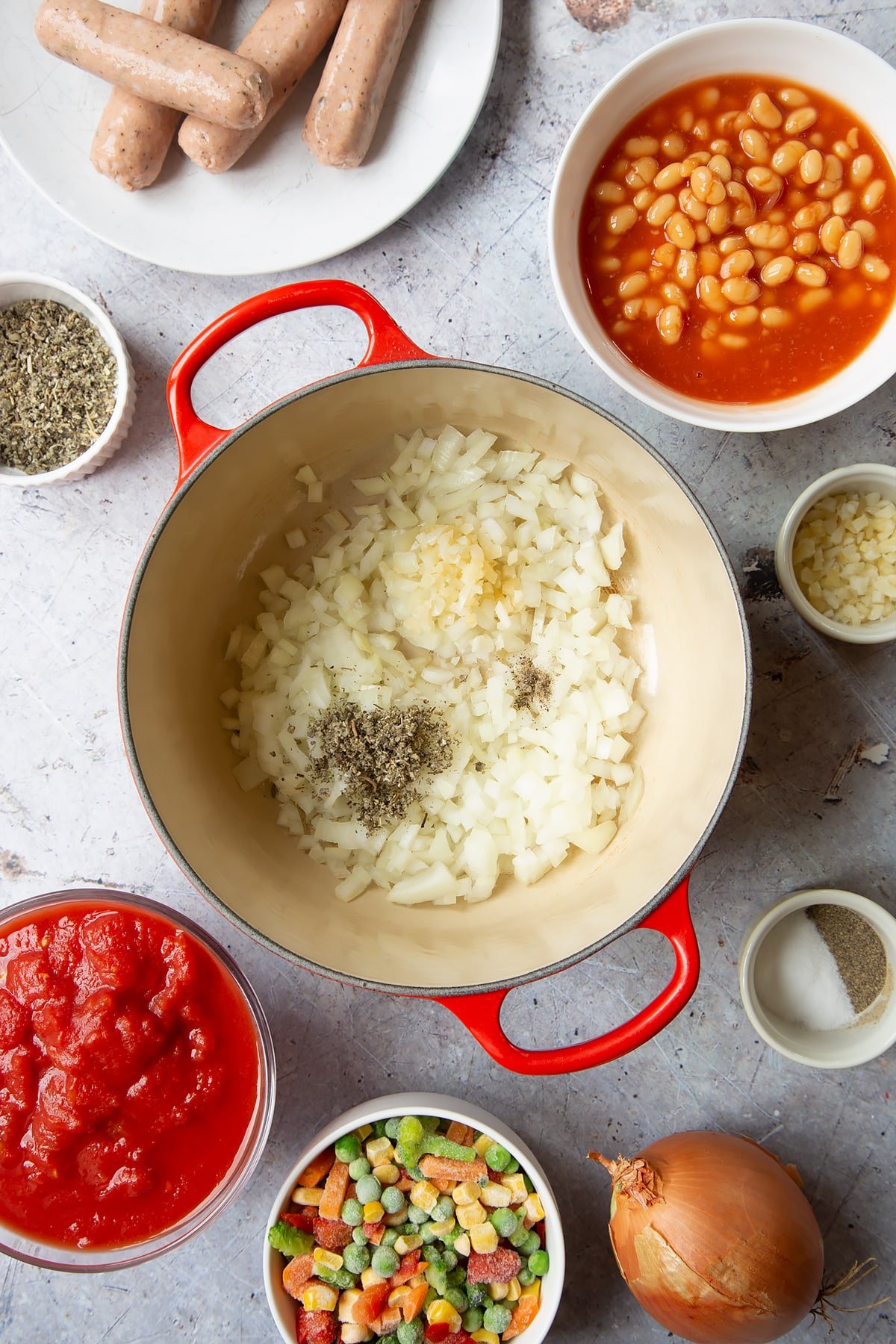 Onions, garlic and oregano in a large pot. Ingredients to make veggie sausage and bean casserole surround the pot.