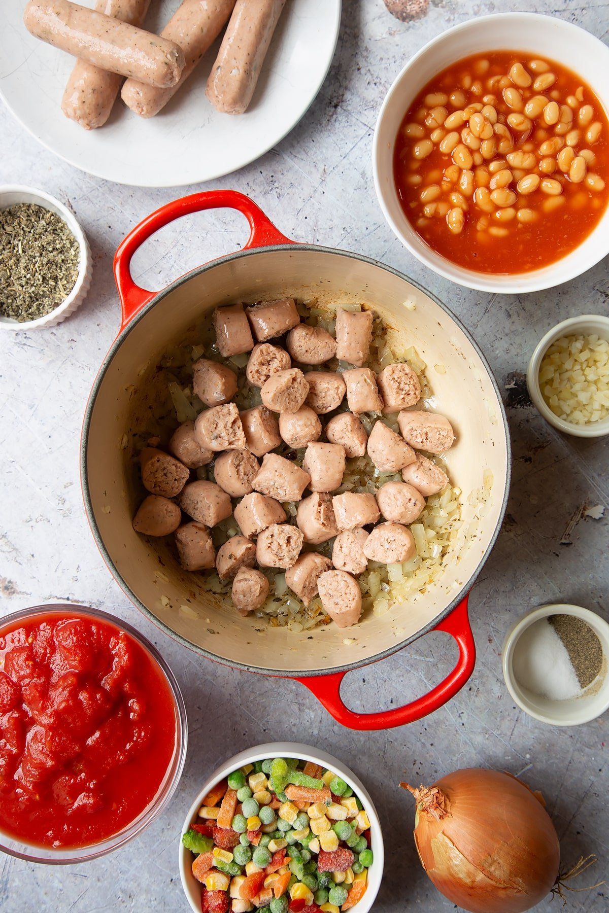 Fried onions in a large pot with chopped vegetarian sausages on top. Ingredients to make veggie sausage and bean casserole surround the pot.