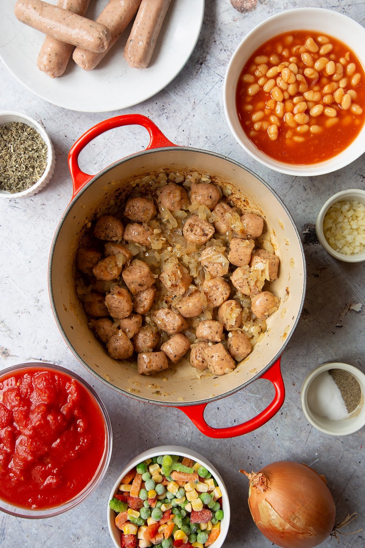 Fried onions and chopped vegetarian sausages in a large pot. Ingredients to make veggie sausage and bean casserole surround the pot.