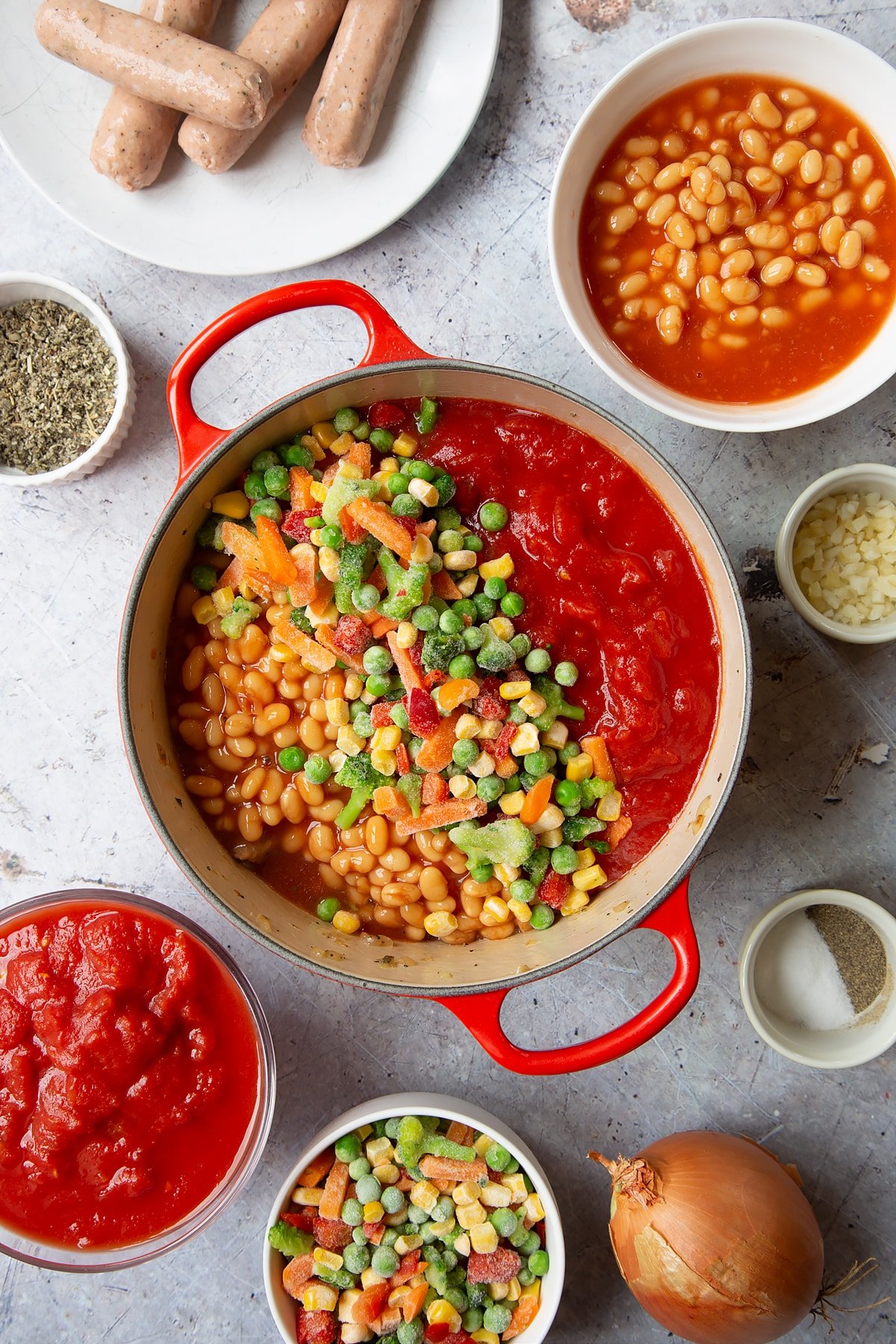 Fried onions and chopped vegetarian sausages in a large pot with beans, chopped tomatoes and frozen chopped vegetables on top. Ingredients to make veggie sausage and bean casserole surround the pot.