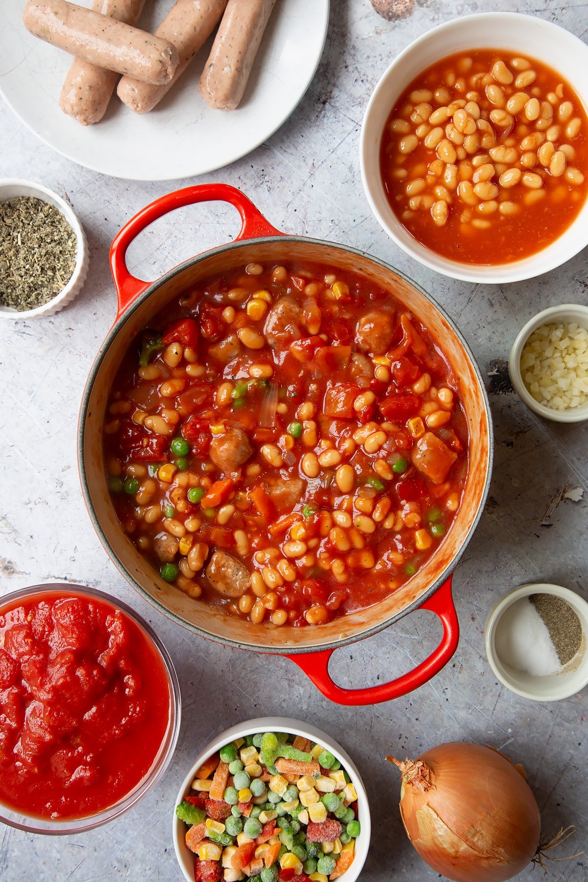 Fried onions, chopped vegetarian sausages, beans, chopped tomatoes and frozen chopped vegetables mixed together in a large pot. Ingredients to make veggie sausage and bean casserole surround the pot.