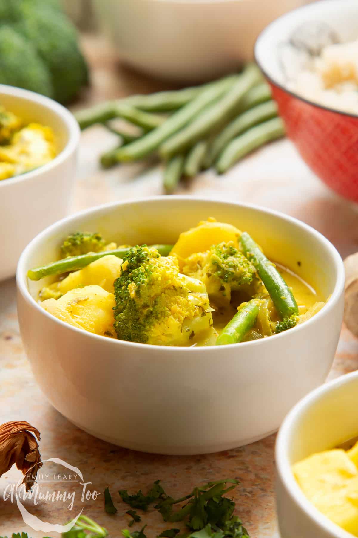 Vegetarian yellow curry with broccoli, potato and green beans, served to small white bowls. A bowl of rice is shown in the background.