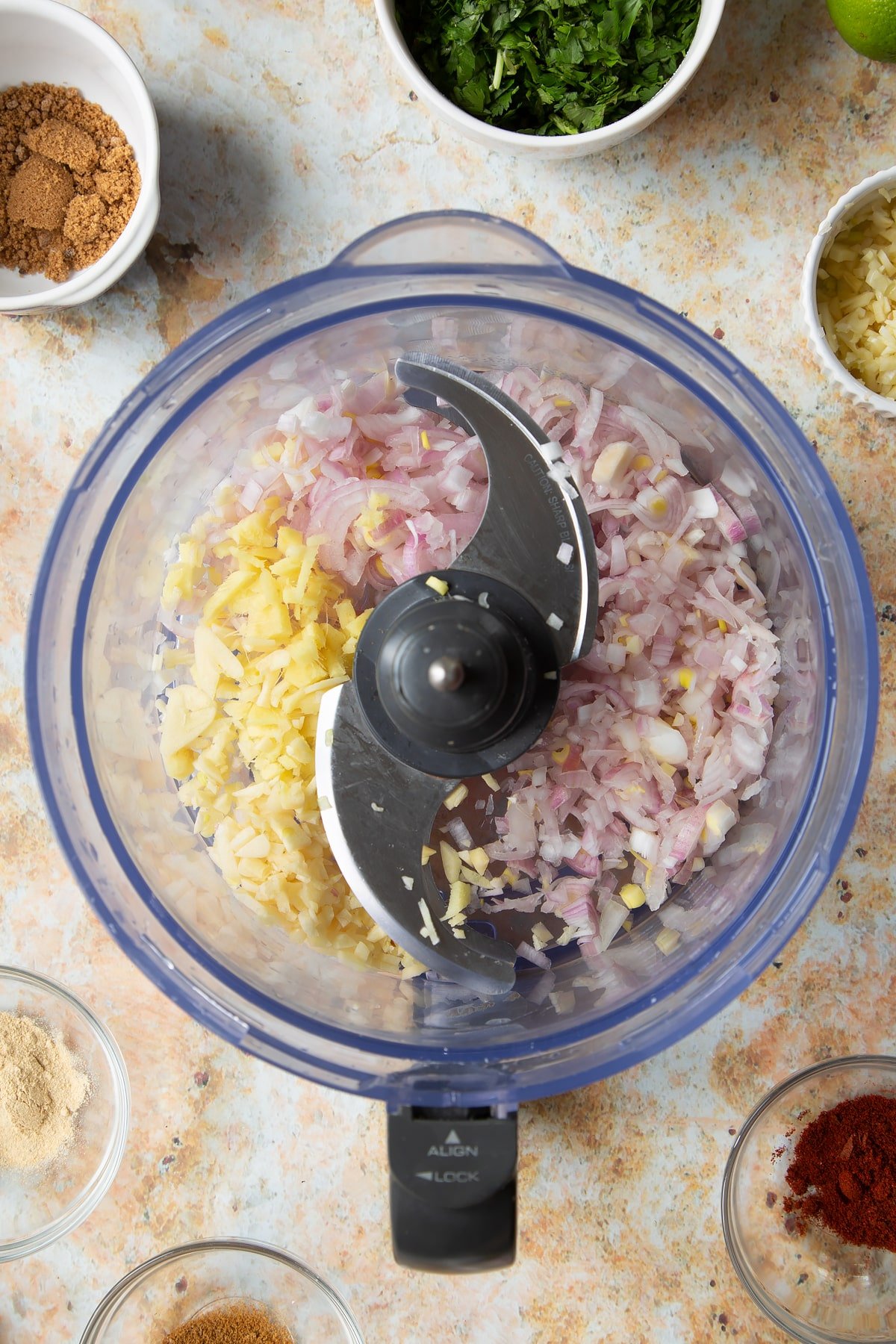 Chopped onion, ginger and garlic in a food processor bowl. Ingredients to make vegetarian yellow curry surround the bowl.