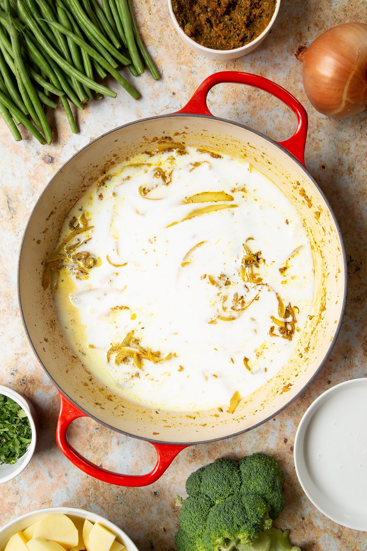 Fried onions, yellow curry paste and coconut milk in a large pan. Ingredients to make vegetarian yellow curry surround the pan.