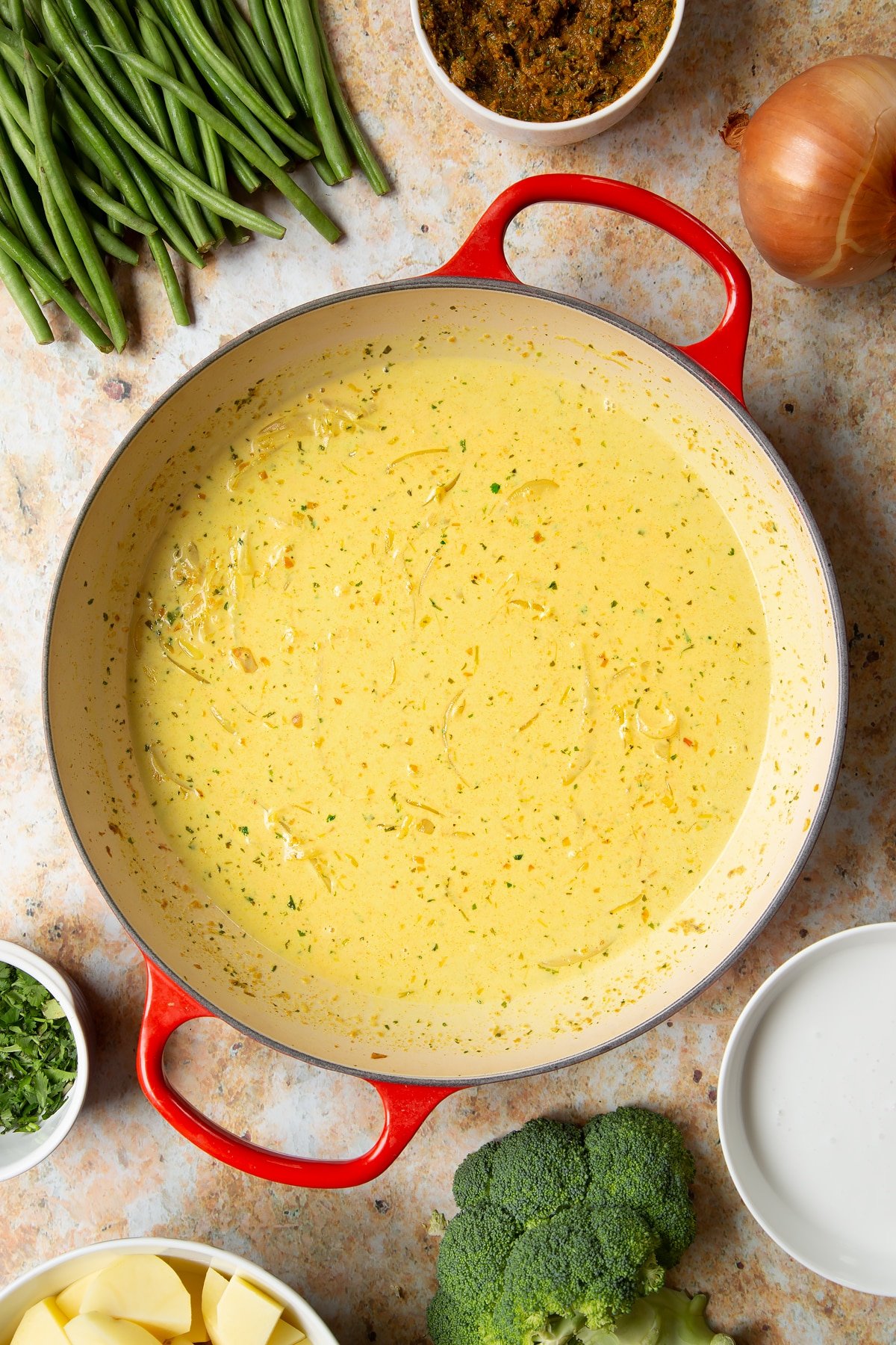 Fried onions, yellow curry paste and coconut milk mixed together in a large pan. Ingredients to make vegetarian yellow curry surround the pan.