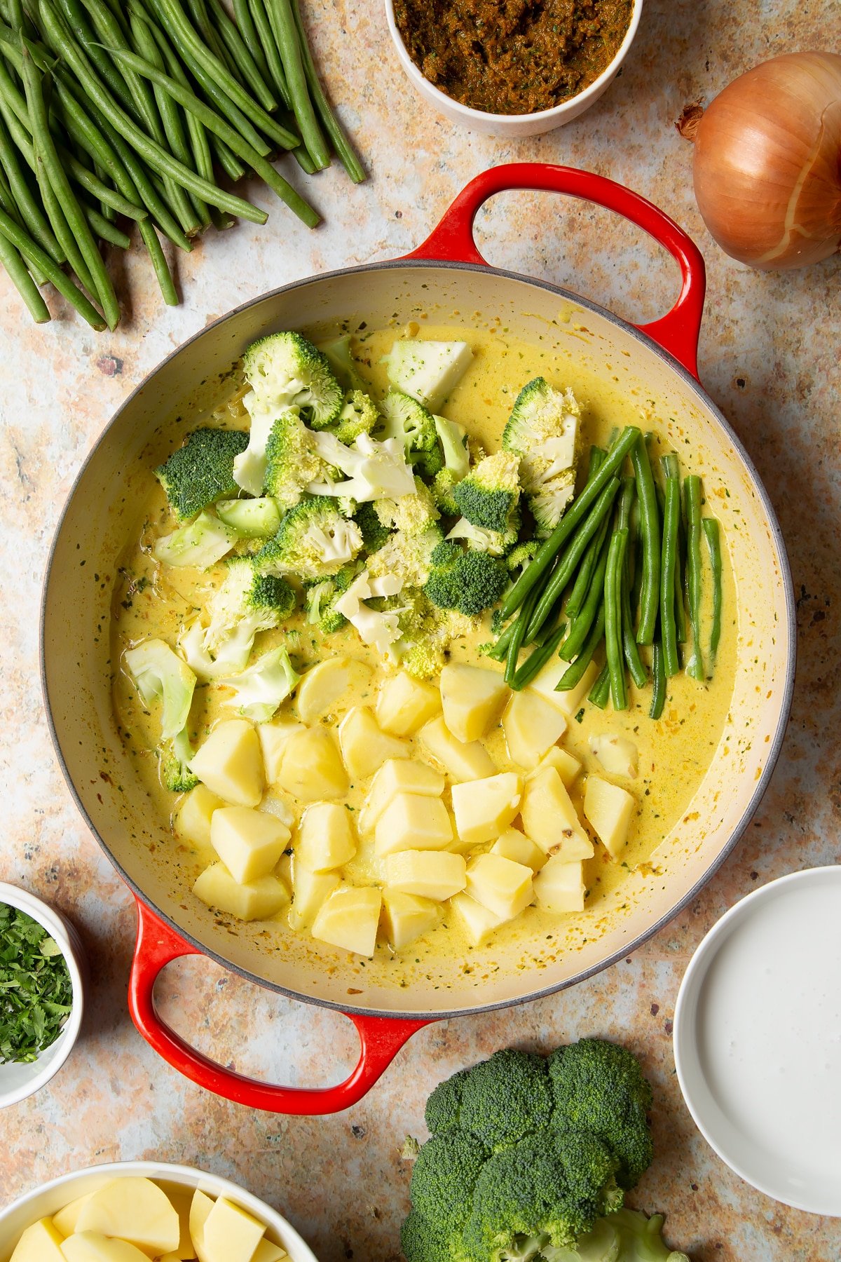 Yellow curry sauce in a large pan with potatoes, green beans and broccoli florets added. Ingredients to make vegetarian yellow curry surround the pan.