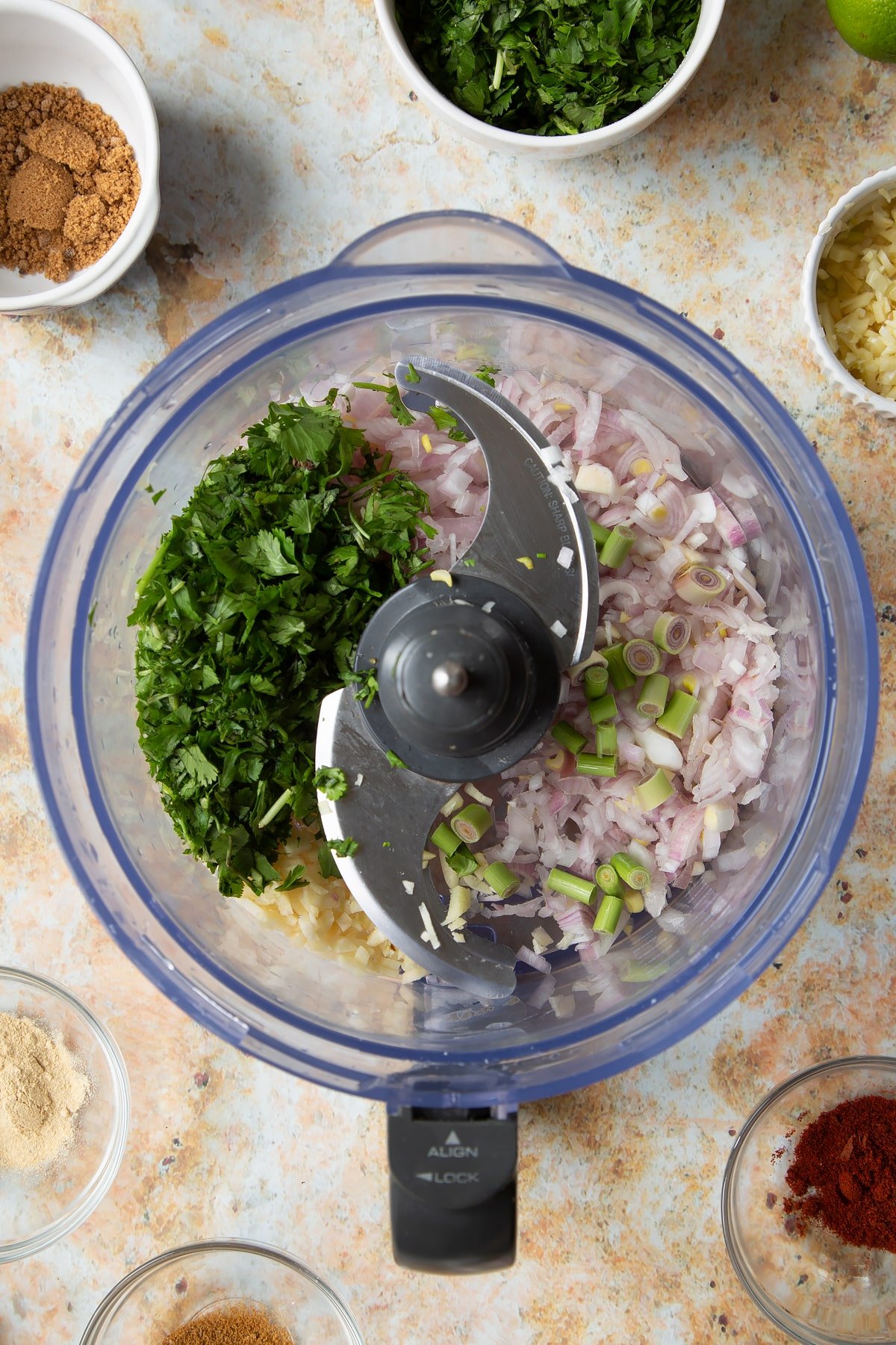 Chopped onion, ginger, garlic, coriander and lemon grass in a food processor bowl. Ingredients to make vegetarian yellow curry surround the bowl.